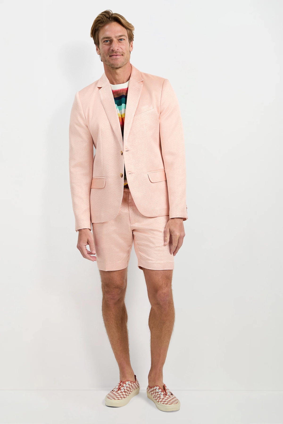A man stands against a white background wearing the Thurston Blazer - Metallic Jacquard with matching shorts, a multicolored striped sweater, and checkered slip-on shoes. He smiles and keeps one hand by his side.