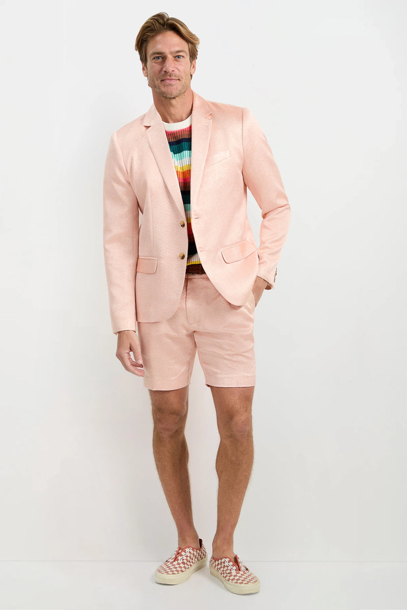 A man stands against a white background wearing the Thurston Blazer - Metallic Jacquard with matching shorts, a colorful striped sweater, and red-and-white checkered slip-ons. He smiles with one hand in his pocket.