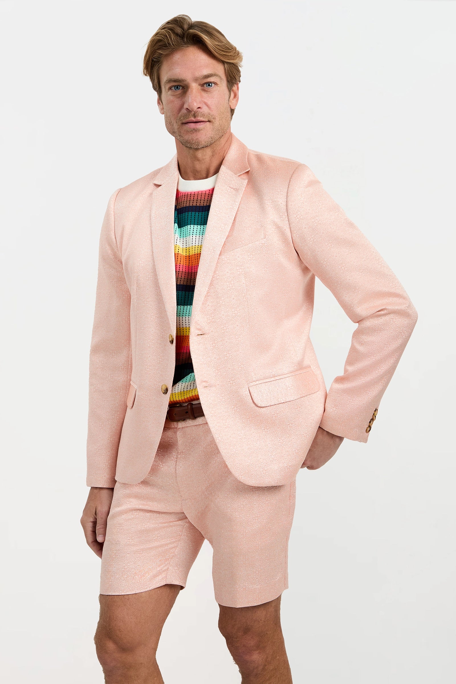 A man poses against a plain background wearing the Thurston Blazer - Metallic Jacquard with matching shorts, his right hand on his hip, and a colorful striped shirt underneath.