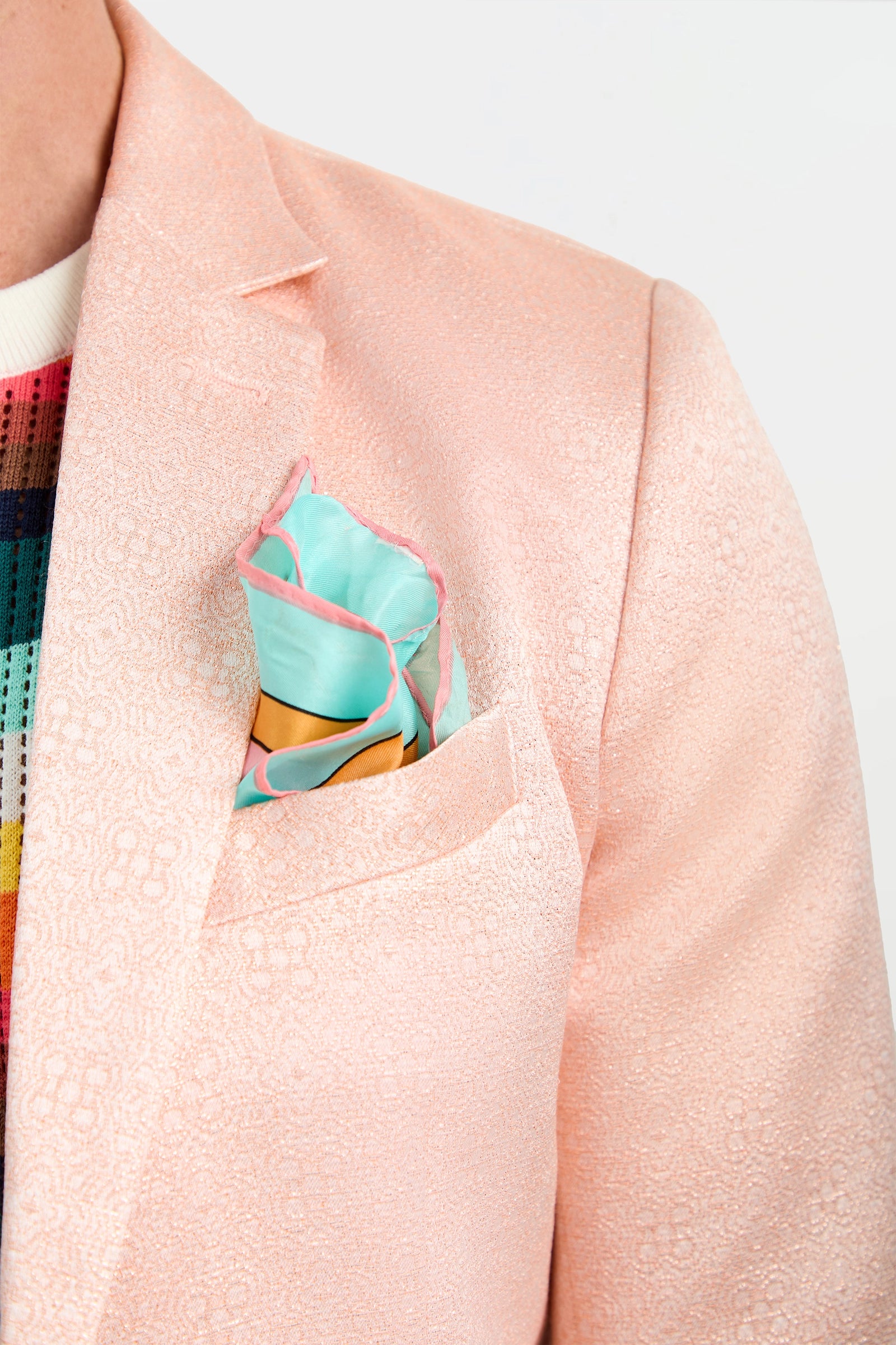 A close-up of a person wearing the Thurston Blazer - Metallic Jacquard, featuring a light pink textured fabric, a colorful patterned pocket square in the breast pocket, and a multicolored checkered shirt underneath.