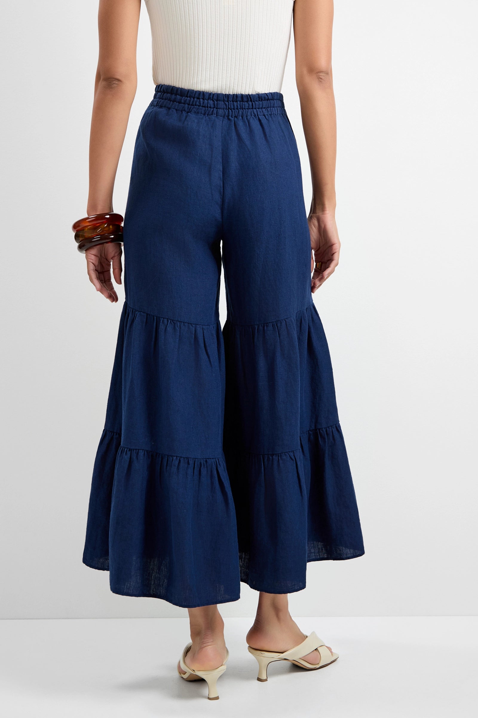 Someone wearing the Chill Out Tiered Wide-Leg Pants in navy blue, paired with a sleeveless cream top, beige heeled sandals, and chunky bangles, is shown from the back against a plain background.