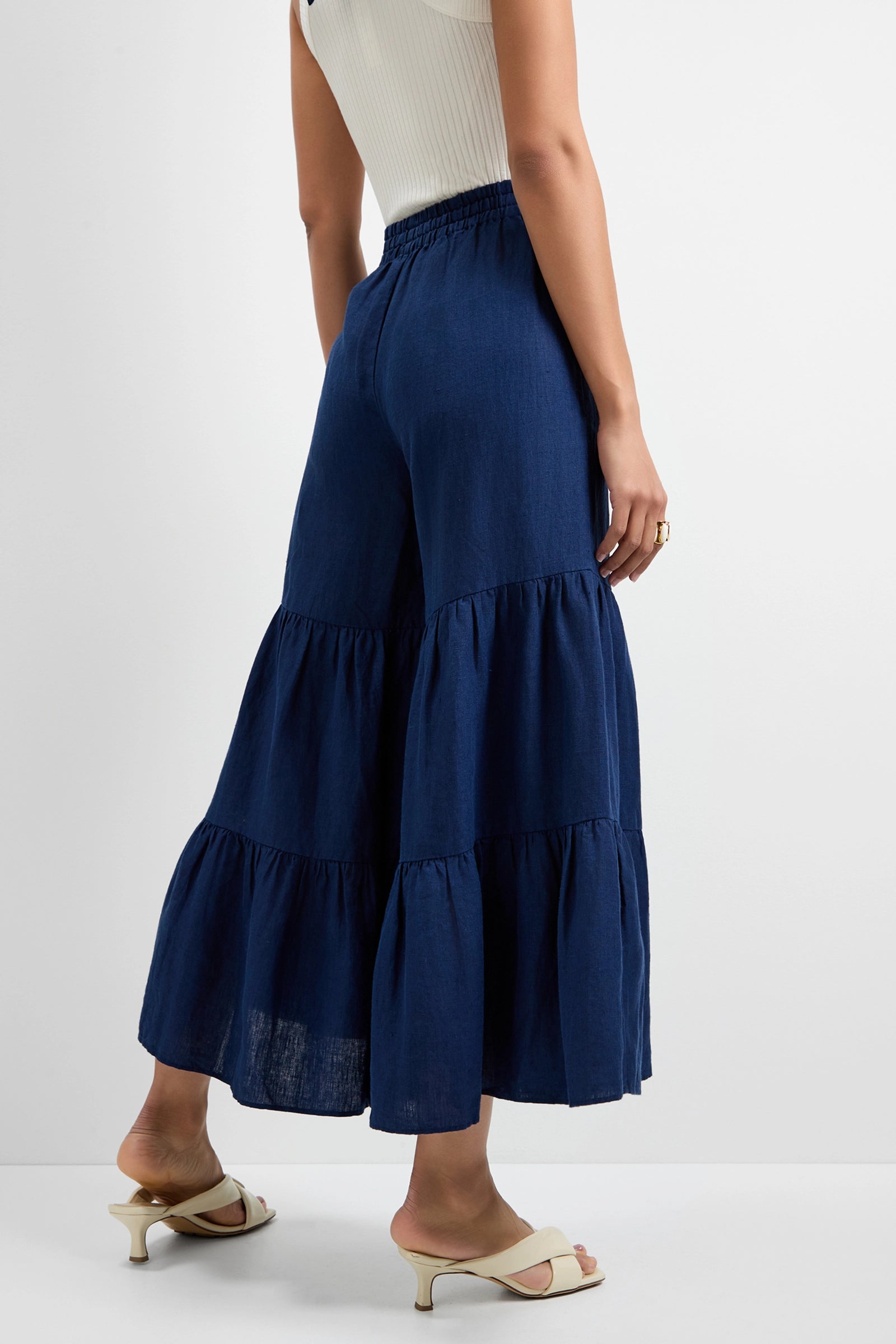 A woman stands against a white wall in a sleeveless ribbed white top, beige heeled sandals, and Chill Out Tiered Wide-Leg Pants in navy blue. Her back is turned with one hand resting at her side.