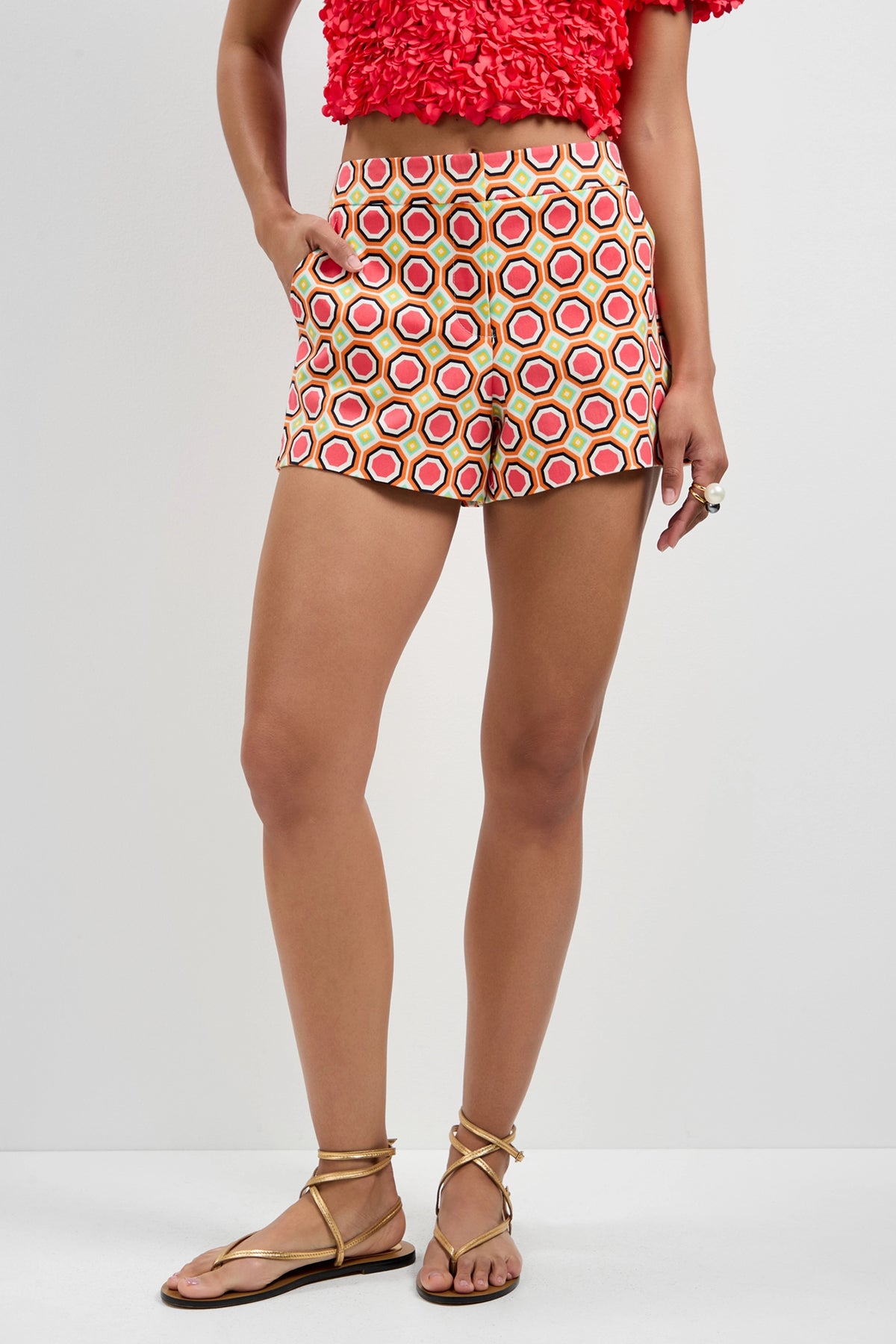 A woman poses against a plain white background wearing the Corbin Shorts - Salerno, featuring a red, orange, and white geometric pattern, paired with a red textured crop top, gold sandals, and a ring.