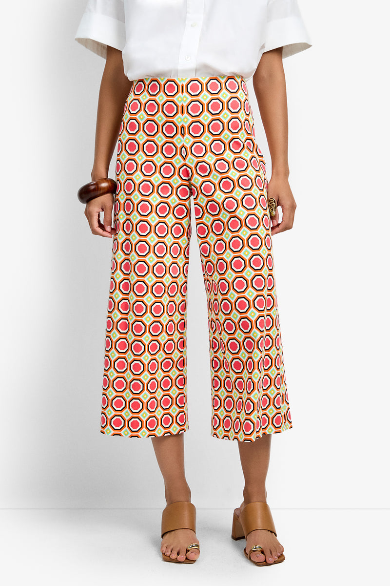Someone wears the Diona Crop Pant—a high-waisted, wide-leg cropped pant with a colorful red, pink, and yellow hexagon pattern—paired with a white top, tan sandals, and a brown bangle. The head is not visible.