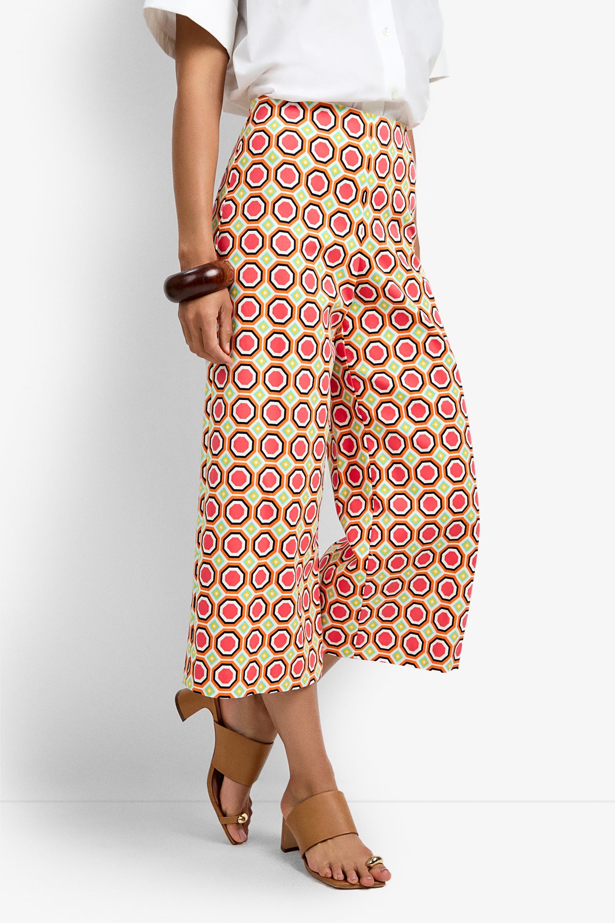 Only the lower half of a person is visible, showcasing the Diona Crop Pant with bold red, yellow, and white circular patterns, paired with tan heeled sandals and a chunky brown bracelet.
