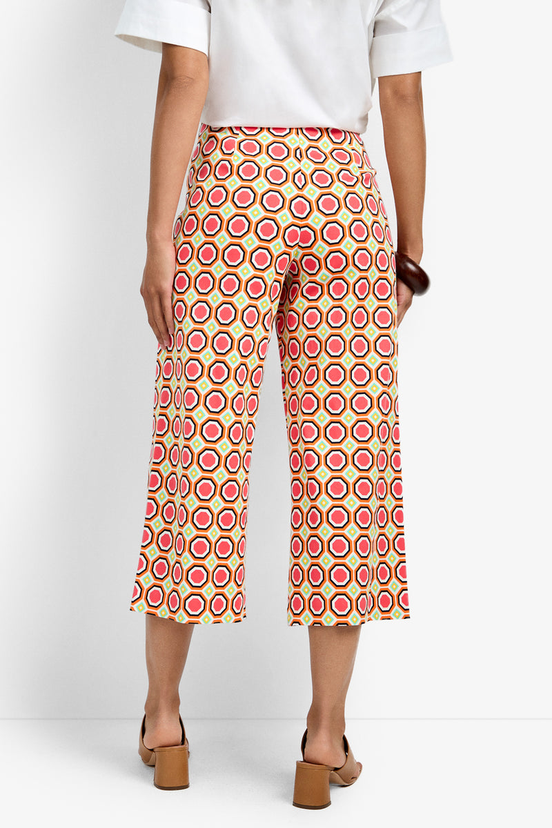 A person stands facing away, wearing the Diona Crop Pant featuring a red, yellow, and white hexagon pattern. They pair it with a white short-sleeve shirt and tan heeled sandals.