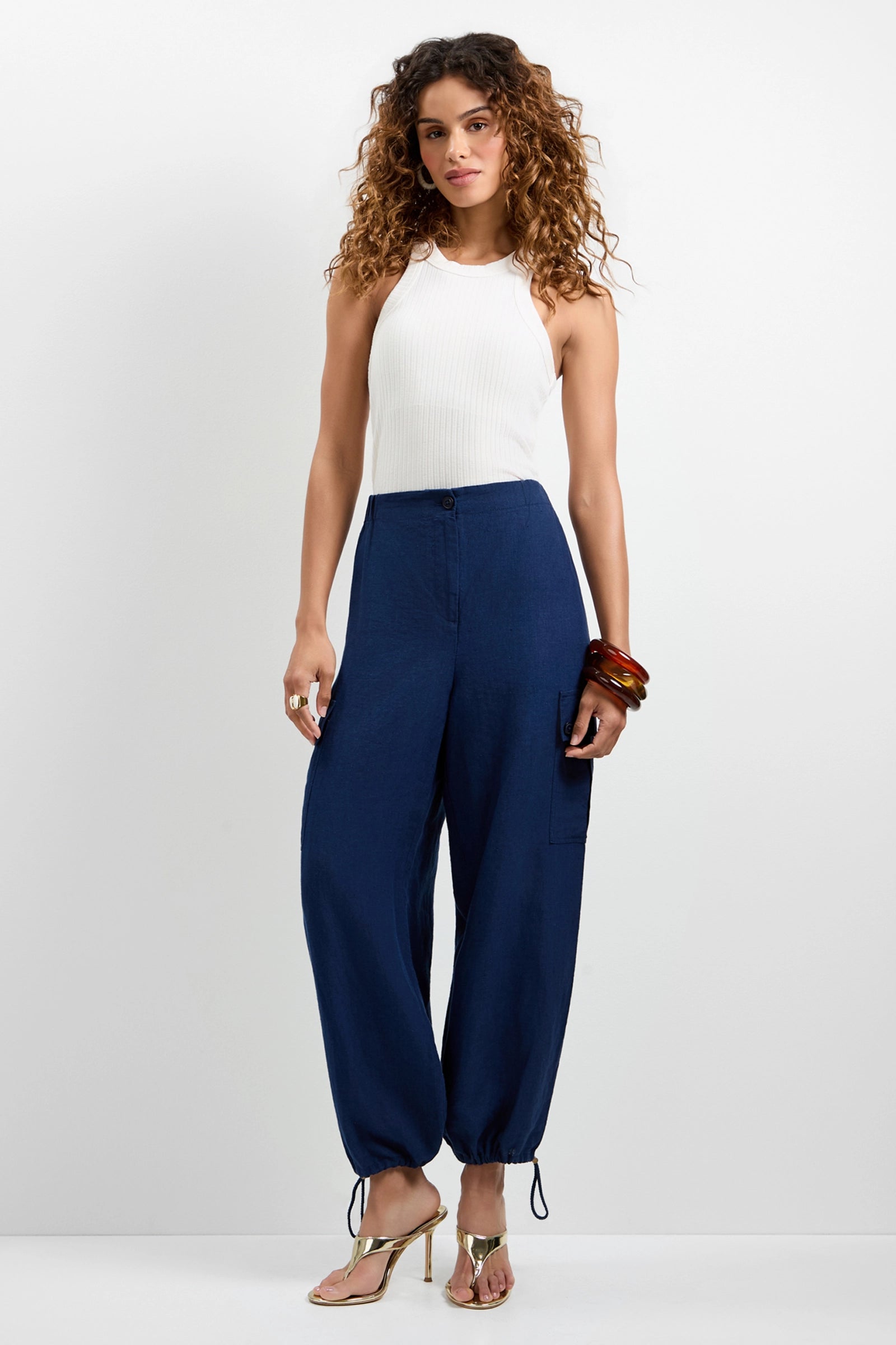 A woman with curly hair wears a white sleeveless top and Alico Cargo Pants—loose navy blue pants with drawstring hems—paired with bangles and heeled sandals, posing against a plain white background.