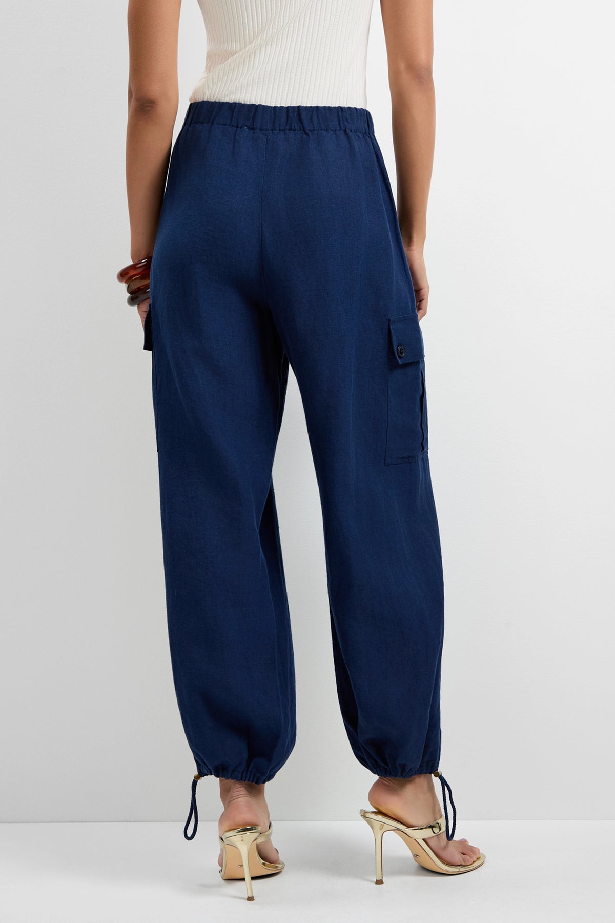 A person wearing Alico Cargo Pants in navy blue with an elastic waistband and drawstring ankles, paired with white high-heeled sandals and a sleeveless top, stands against a plain background.