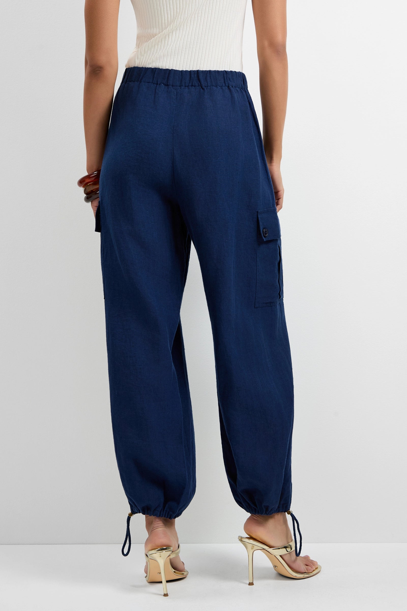 A person wearing Alico Cargo Pants in navy blue with an elastic waistband and drawstring ankles, paired with white high-heeled sandals and a sleeveless top, stands against a plain background.