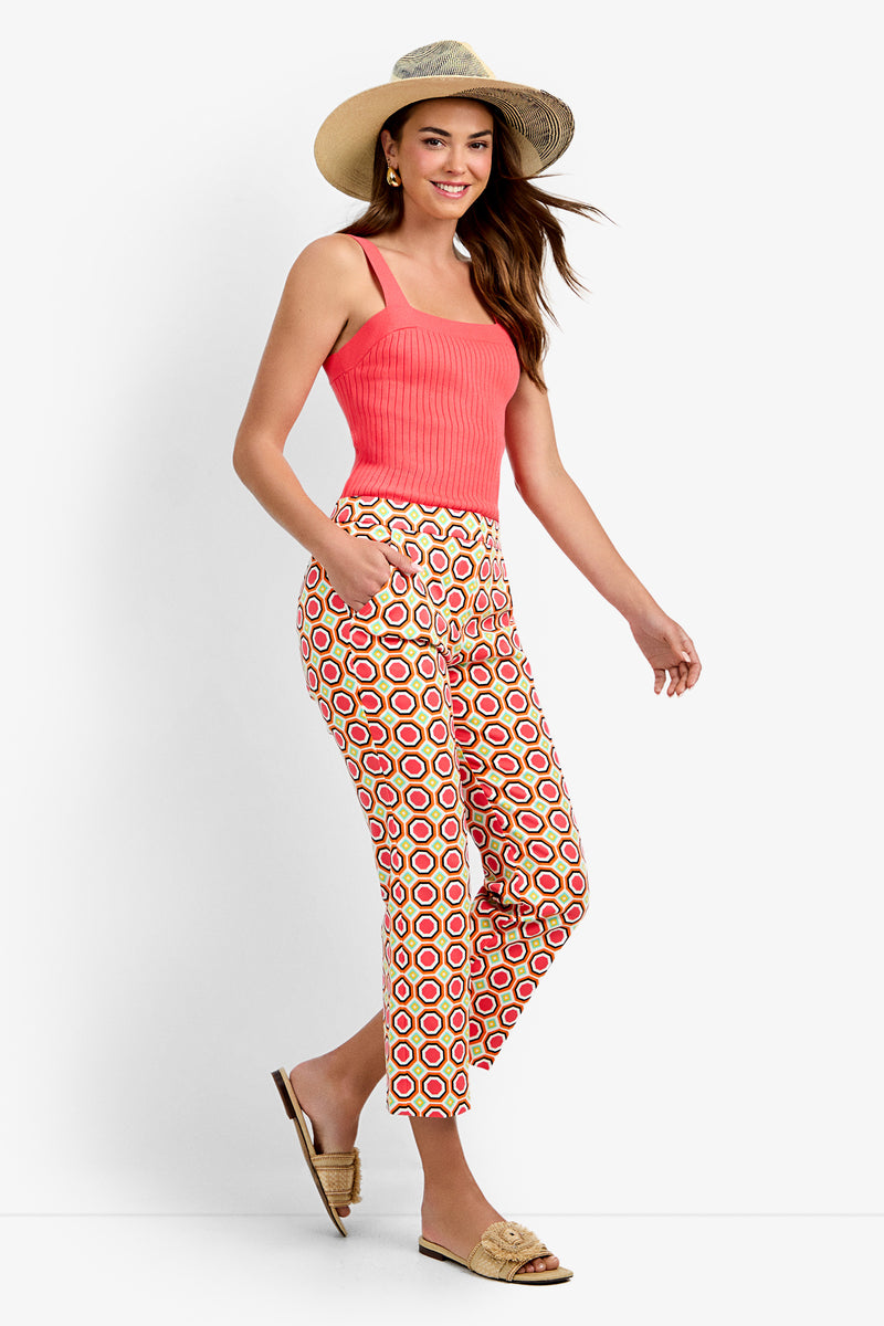 A woman stands smiling with hands in her pockets, wearing Lulu Cropped Pants - Salerno, a coral tank top, a straw hat, and woven slip-on sandals against a white background.