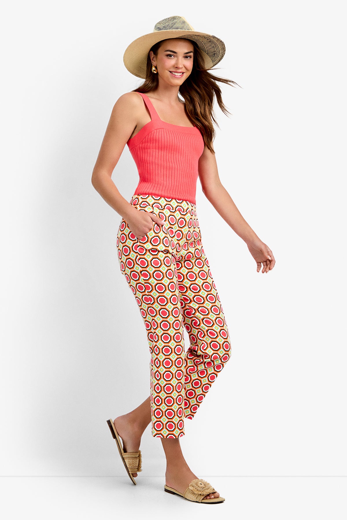 A woman stands smiling with hands in her pockets, wearing Lulu Cropped Pants - Salerno, a coral tank top, a straw hat, and woven slip-on sandals against a white background.