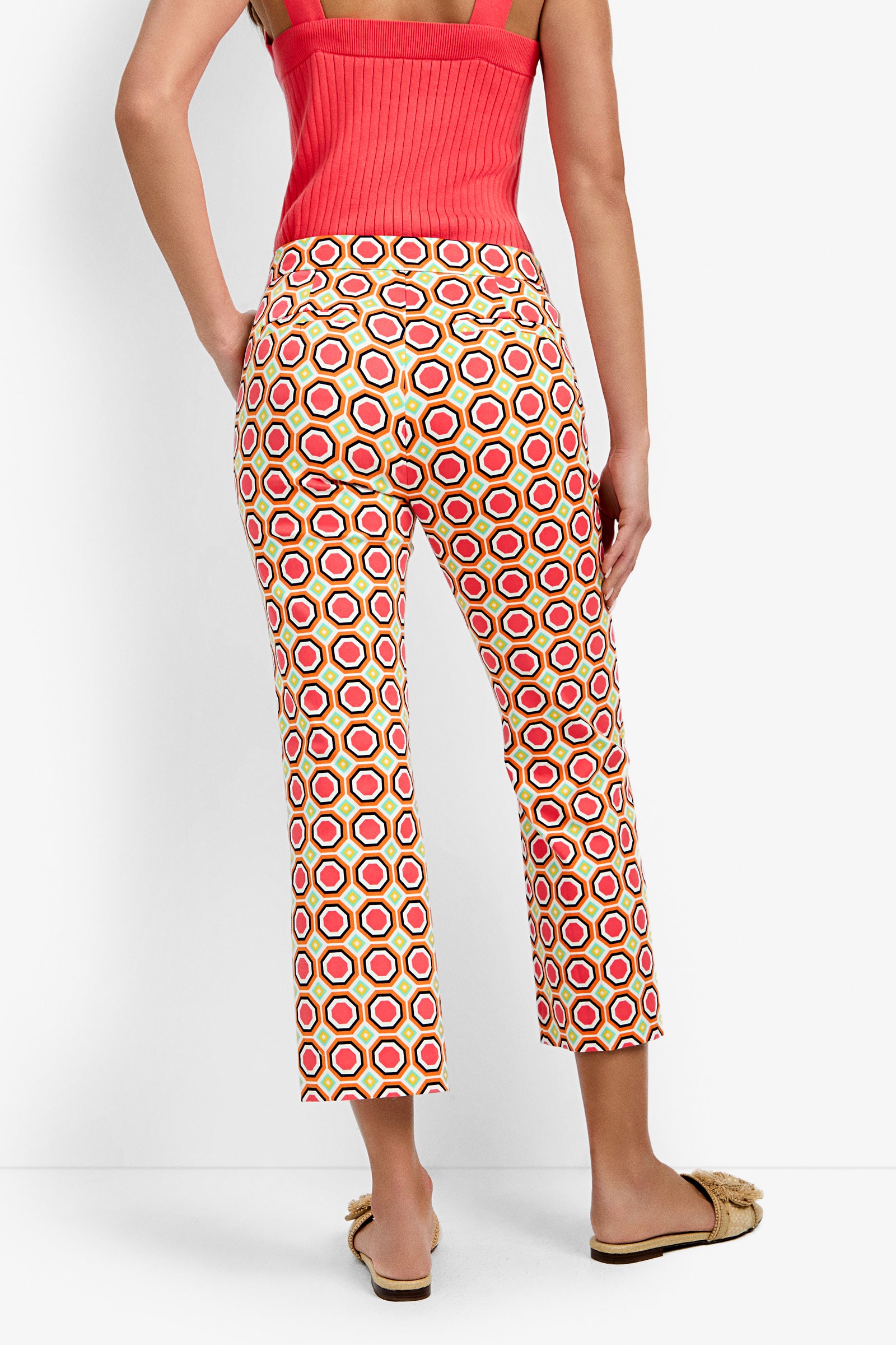 A woman faces away, wearing Lulu Cropped Pants - Salerno with a bold red, yellow, and white geometric pattern, a sleeveless red top, and fluffy slip-on sandals.