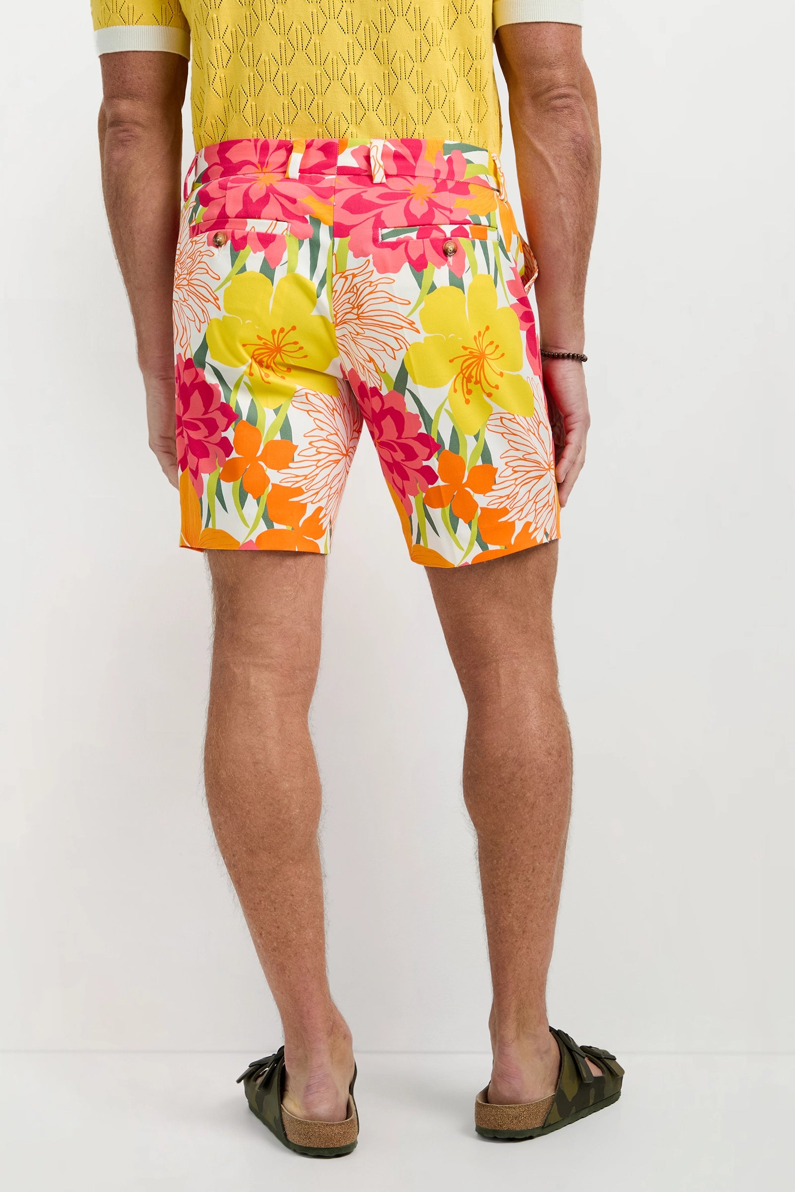 A man stands facing away, wearing the Lawrence Short - Como Herringbone with a yellow patterned shirt and brown sandals. The shorts feature a vibrant floral design with pink, red, yellow, and orange flowers against a plain light background.
