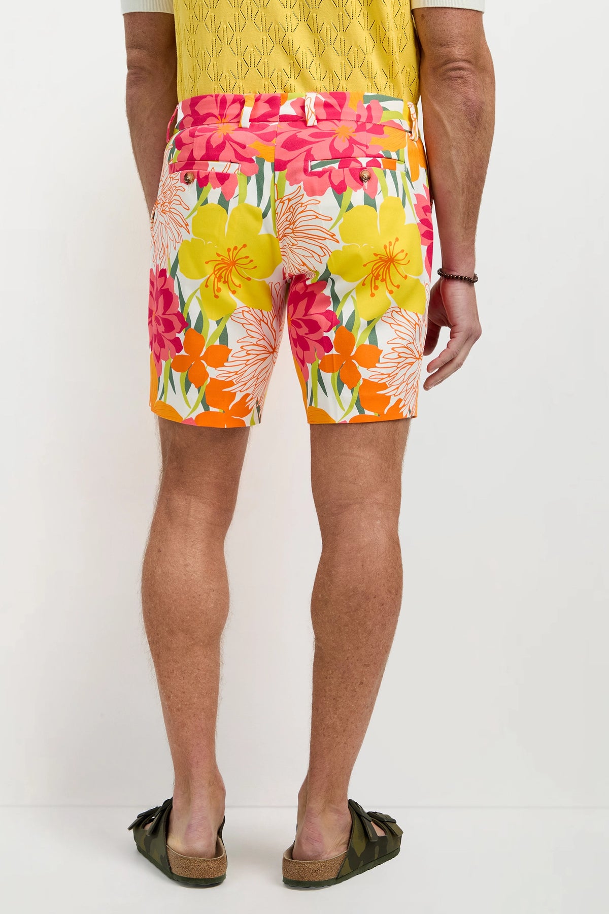 A man stands facing away, wearing the Lawrence Short - Como Herringbone with a yellow patterned shirt, green sandals, and a black bracelet on his left wrist.