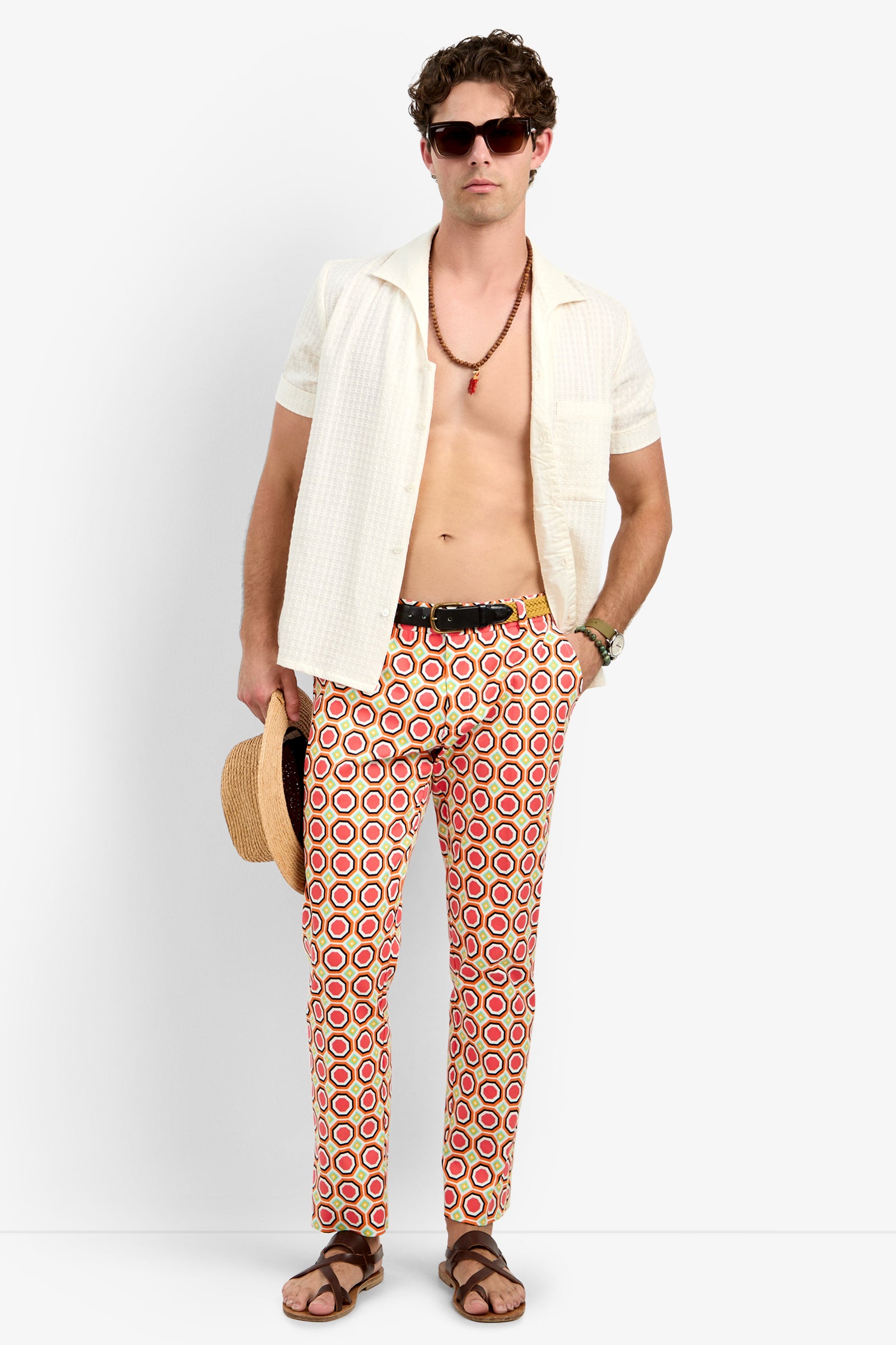 A man in Clyde Slim Trouser - Salerno Herringbone, dark sunglasses, cream short-sleeve shirt, brown sandals, with a straw hat, necklace, and watch stands against a plain white background.