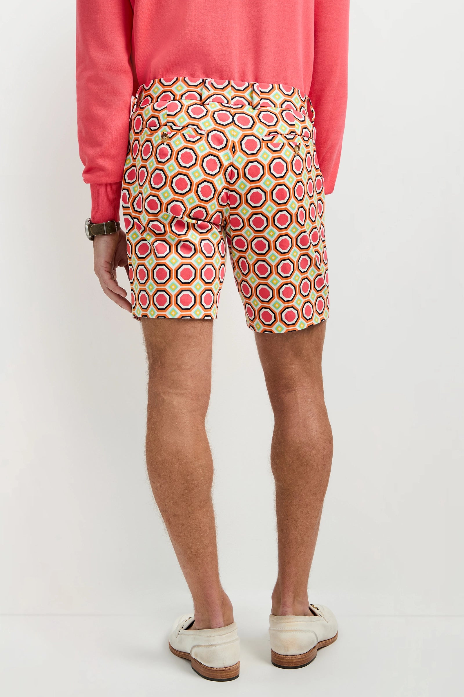 A man faces away, wearing a long-sleeve pink shirt, Lawrence Short - Salerno Herringbone with a red, yellow, and white geometric pattern, a watch, and white loafers against a plain white background.