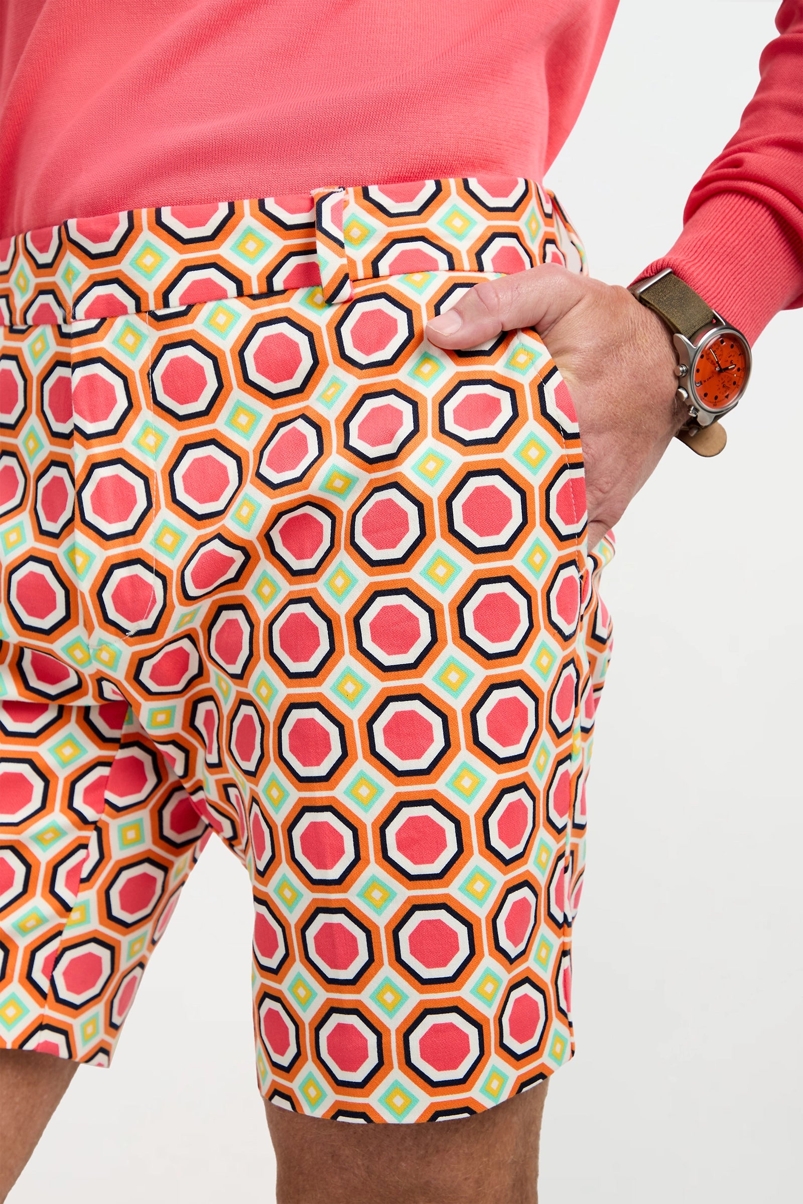 A person wearing Lawrence Short - Salerno Herringbone, a pink shirt, and a brown watch with a red face stands with their hand in their shorts pocket.