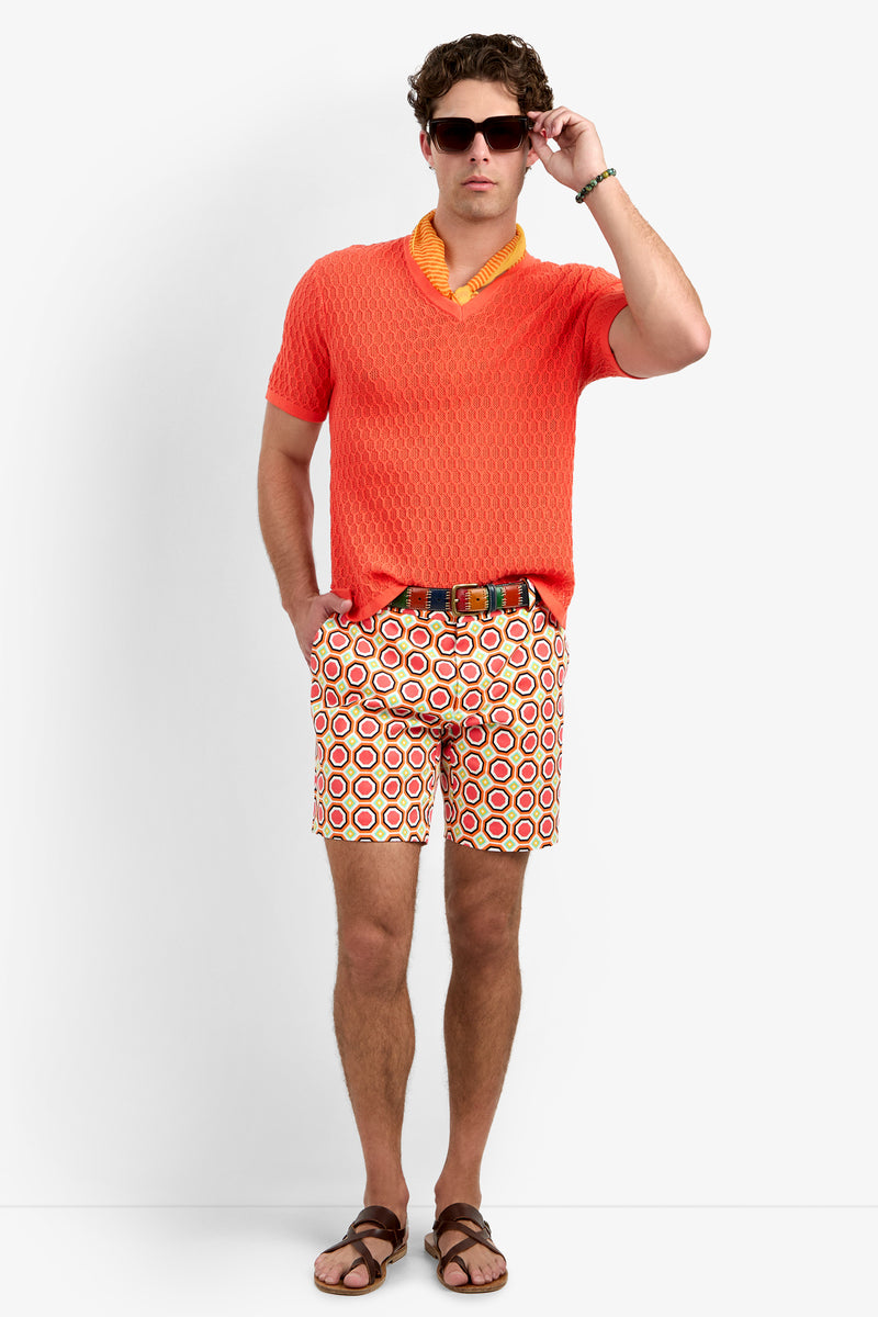 A man with short brown hair stands against a plain background, slightly adjusting his dark sunglasses while wearing an orange textured shirt, brown sandals, and the Lawrence Short - Salerno Herringbone with a belt.