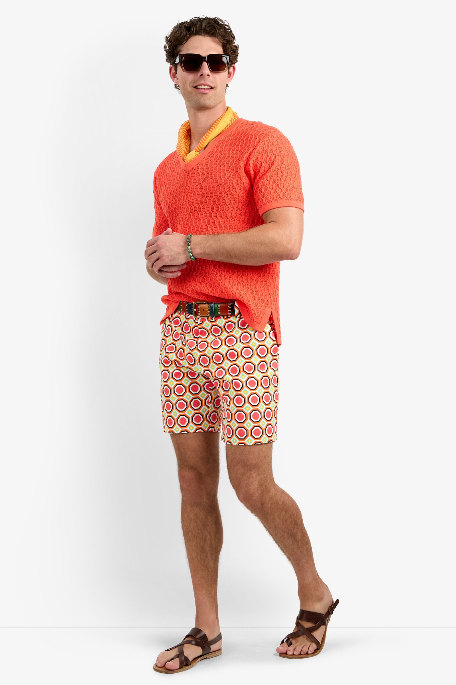 A man stands confidently against a plain white background, wearing sunglasses, an orange shirt with a yellow collar, the Lawrence Short - Salerno Herringbone, a belt, and brown sandals.