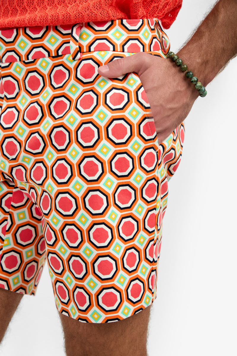 A person wearing the Lawrence Short - Salerno Herringbone, featuring colorful geometric patterns, paired with a coral shirt and a green beaded bracelet, stands with one hand in their pocket.