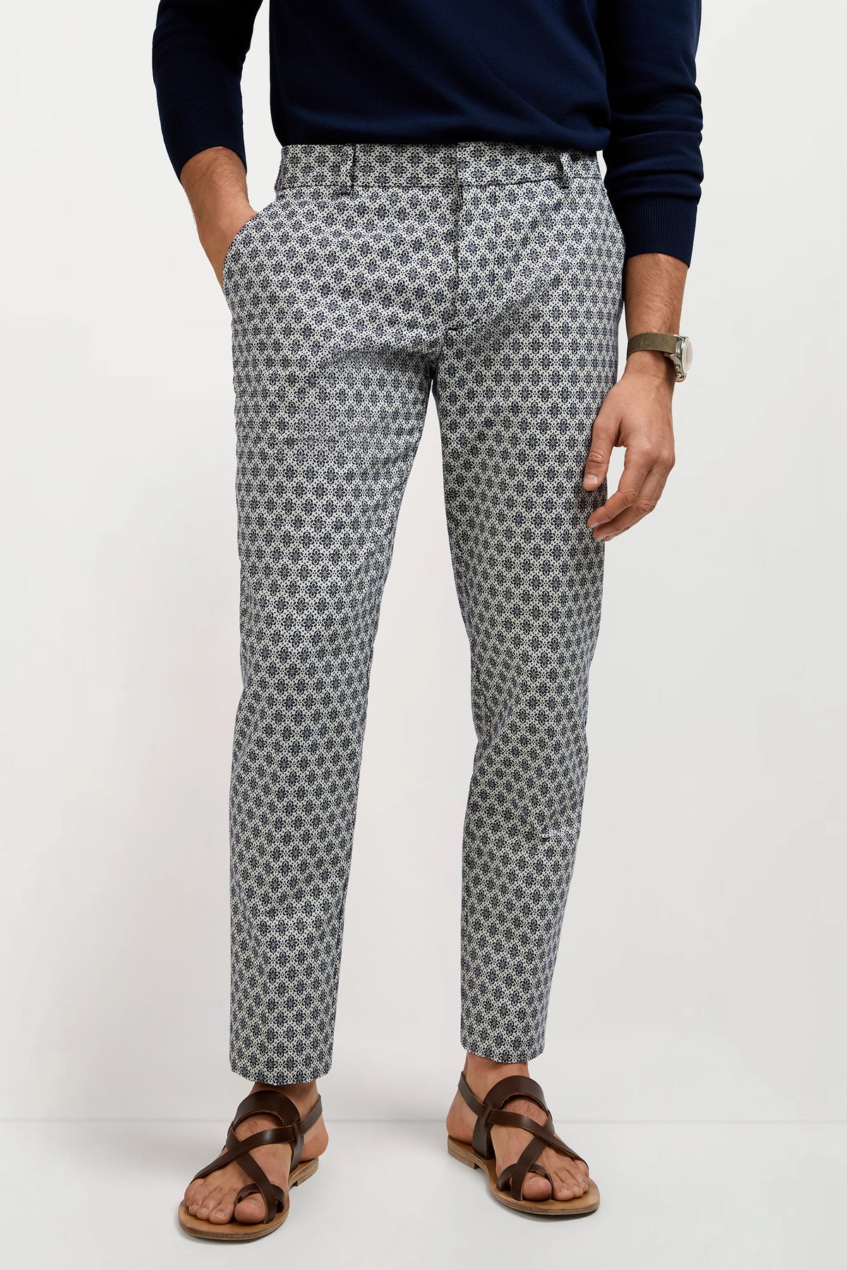 A man wears the Clyde Slim Trouser - Daisy Jacquard, featuring a patterned blue and white design, paired with a dark blue sweater and brown sandals, standing with one hand in his pocket against a plain backdrop.