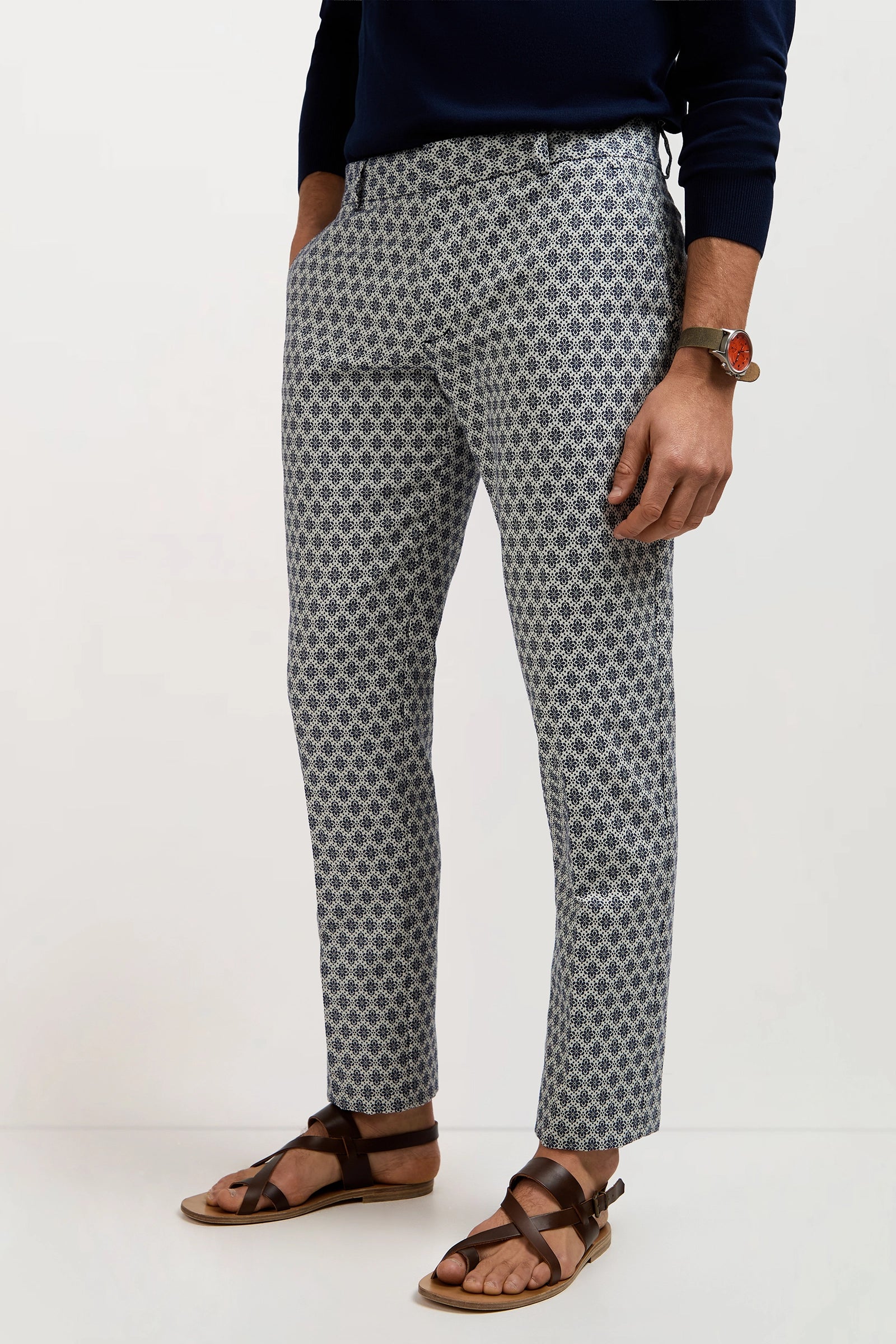 A person models the Clyde Slim Trouser - Daisy Jacquard, paired with a navy sweater, brown leather sandals, and a brown wristwatch against a plain white background, hands in their pockets.