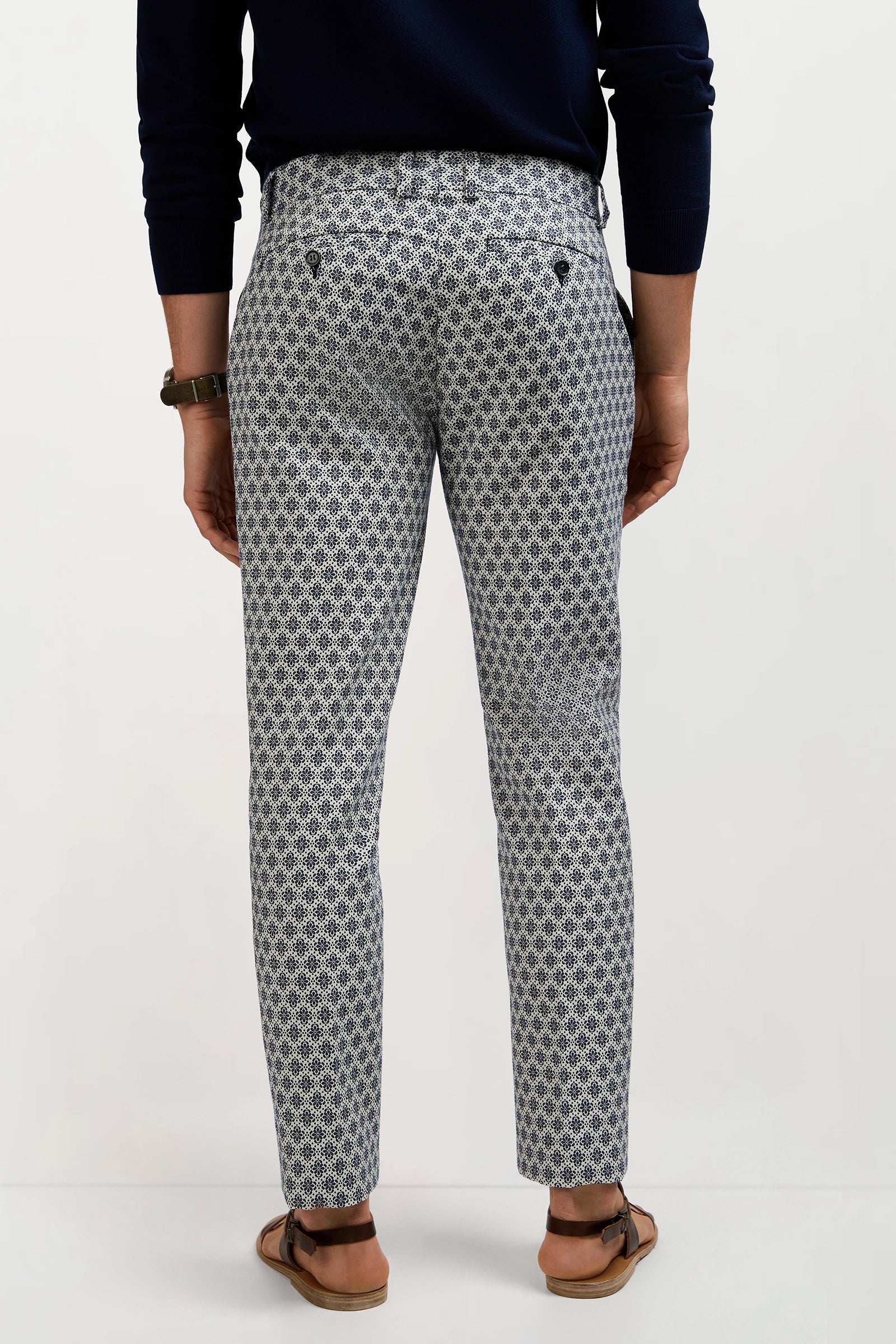 A person stands facing away, wearing the Clyde Slim Trouser - Daisy Jacquard with a dark long-sleeve top, sandals, and a watch against a plain light background.