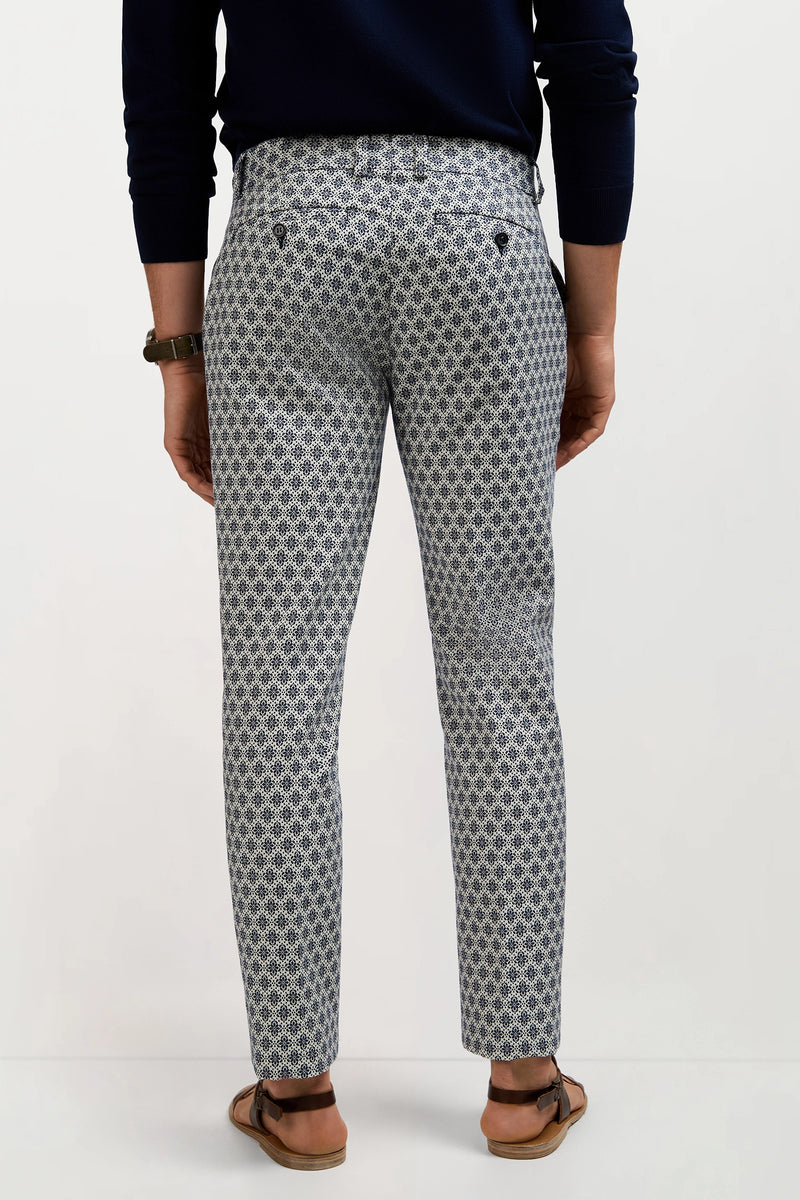 A person stands facing away, wearing the Clyde Slim Trouser - Daisy Jacquard with a dark long-sleeve top, sandals, and a watch against a plain light background.
