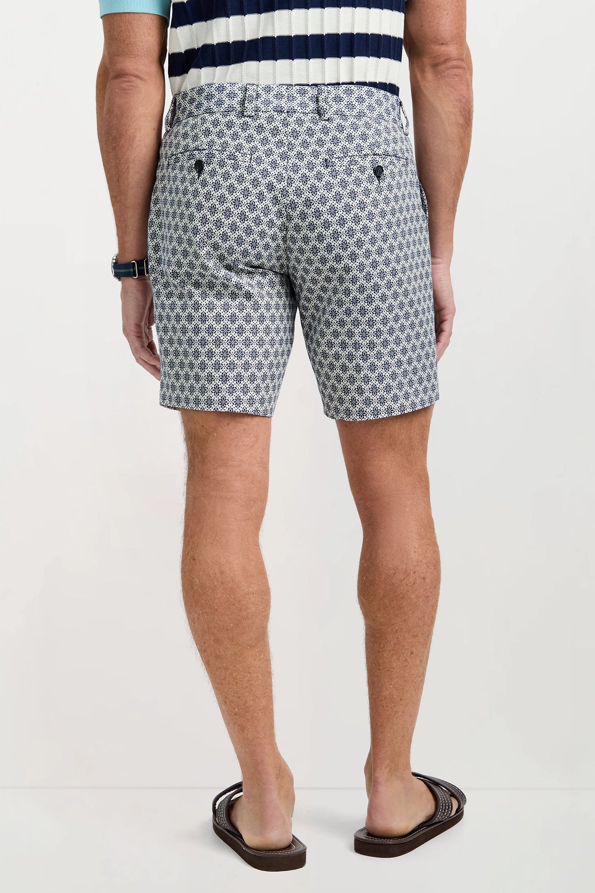 A man stands with his back to the camera, wearing the Lawrence Short - Daisy Jacquard, a patterned pair of shorts, styled with a striped short-sleeve shirt, flip-flops, and a watch against a plain background.