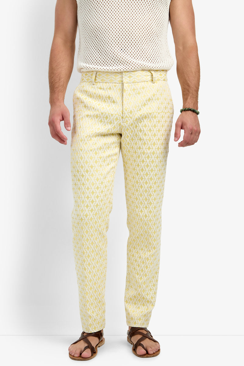 A man wearing the Clyde Slim Trouser - Crossroads Jacquard, a white sleeveless mesh top, brown sandals, and a beaded bracelet stands against a plain white background. His head is not visible in the image.