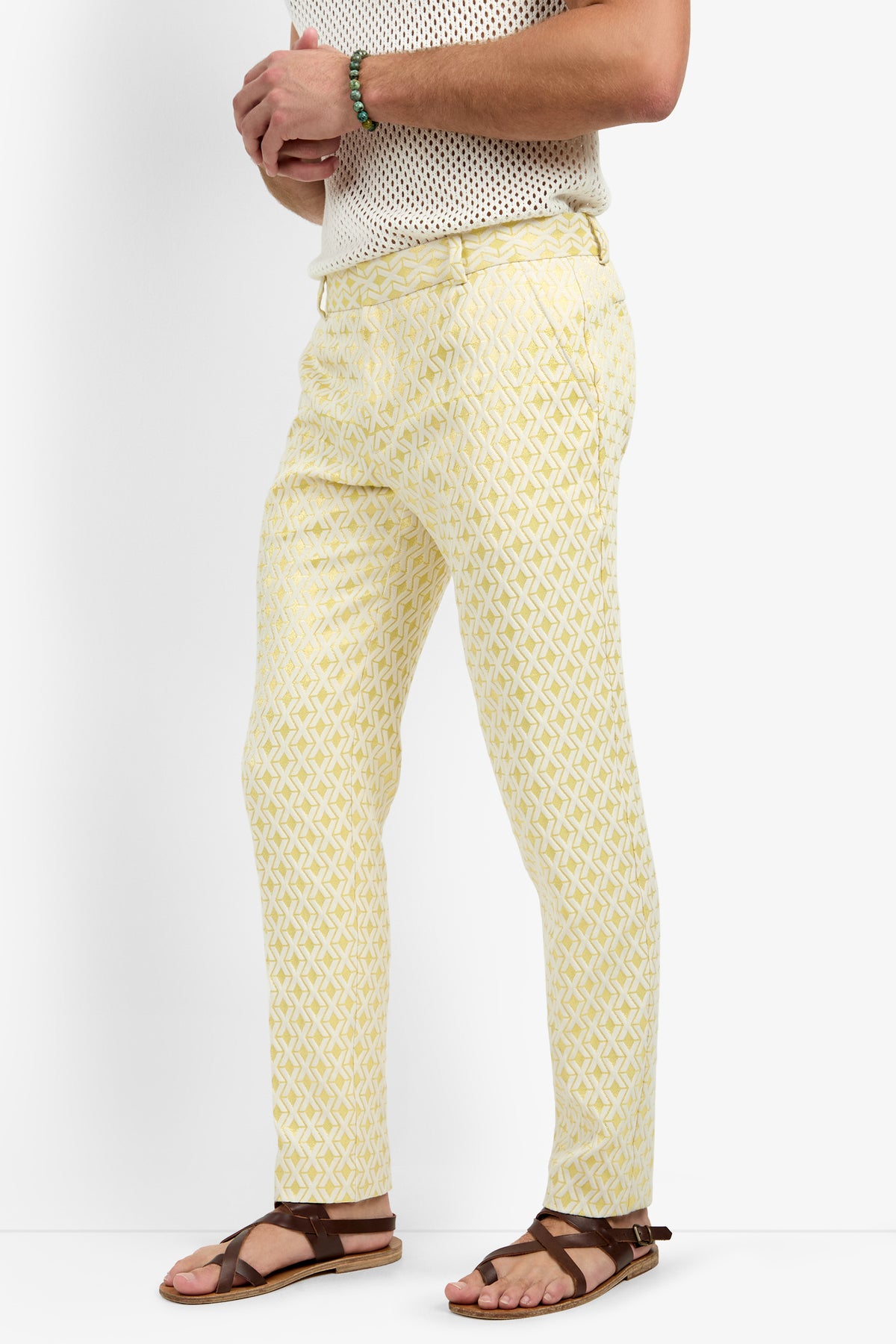 A person wearing the Clyde Slim Trouser - Crossroads Jacquard, a white short-sleeve mesh top, brown sandals, and a green beaded bracelet stands against a white background.