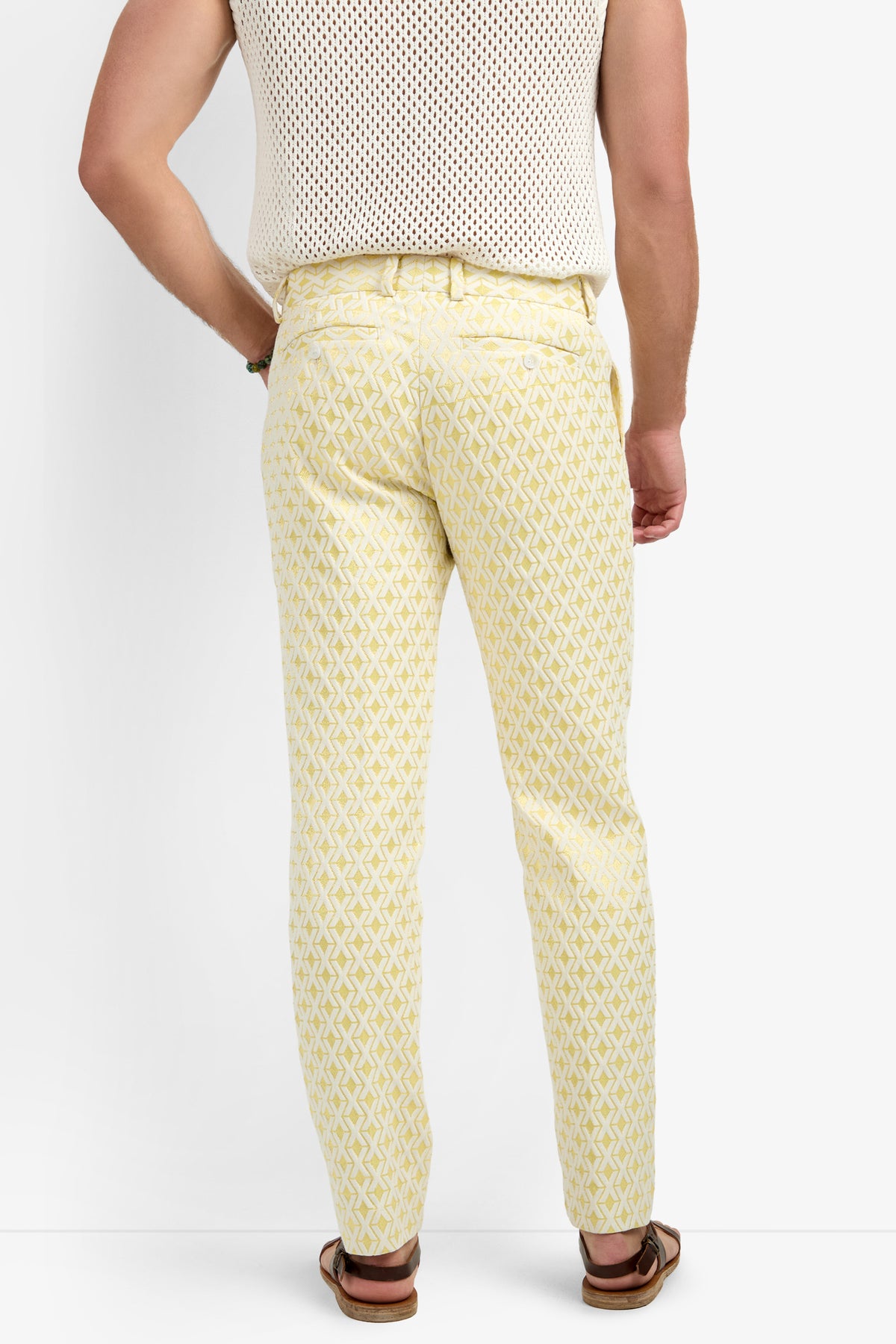 A person stands facing away, wearing the Clyde Slim Trouser in Crossroads Jacquard with a white sleeveless mesh top and brown sandals against a plain white background.