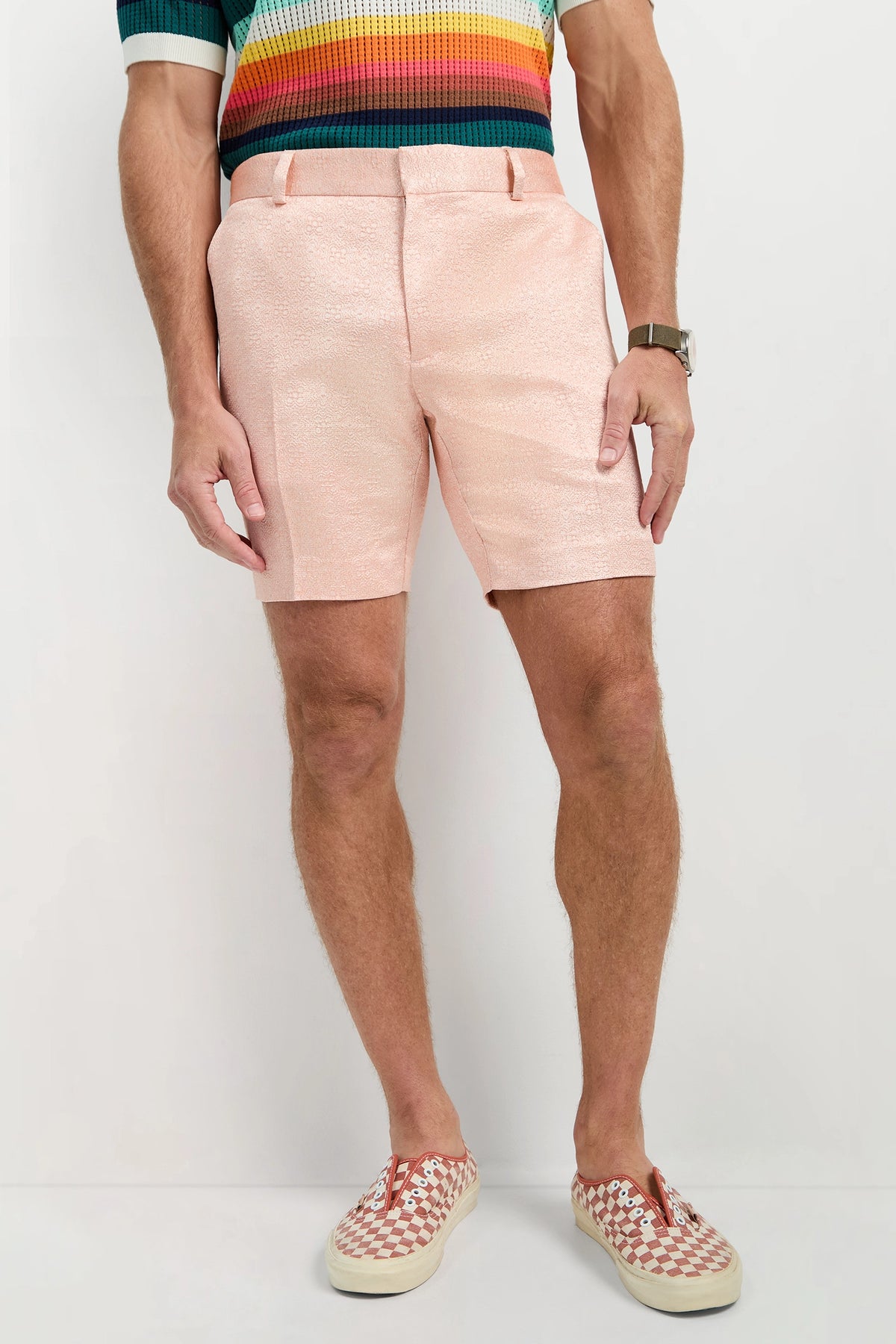 Wearing the Clyde Short - Metallic Jacquard in pale pink with a rainbow-striped shirt, a watch, and red and white checkered slip-on shoes, this person stands against a plain light background.