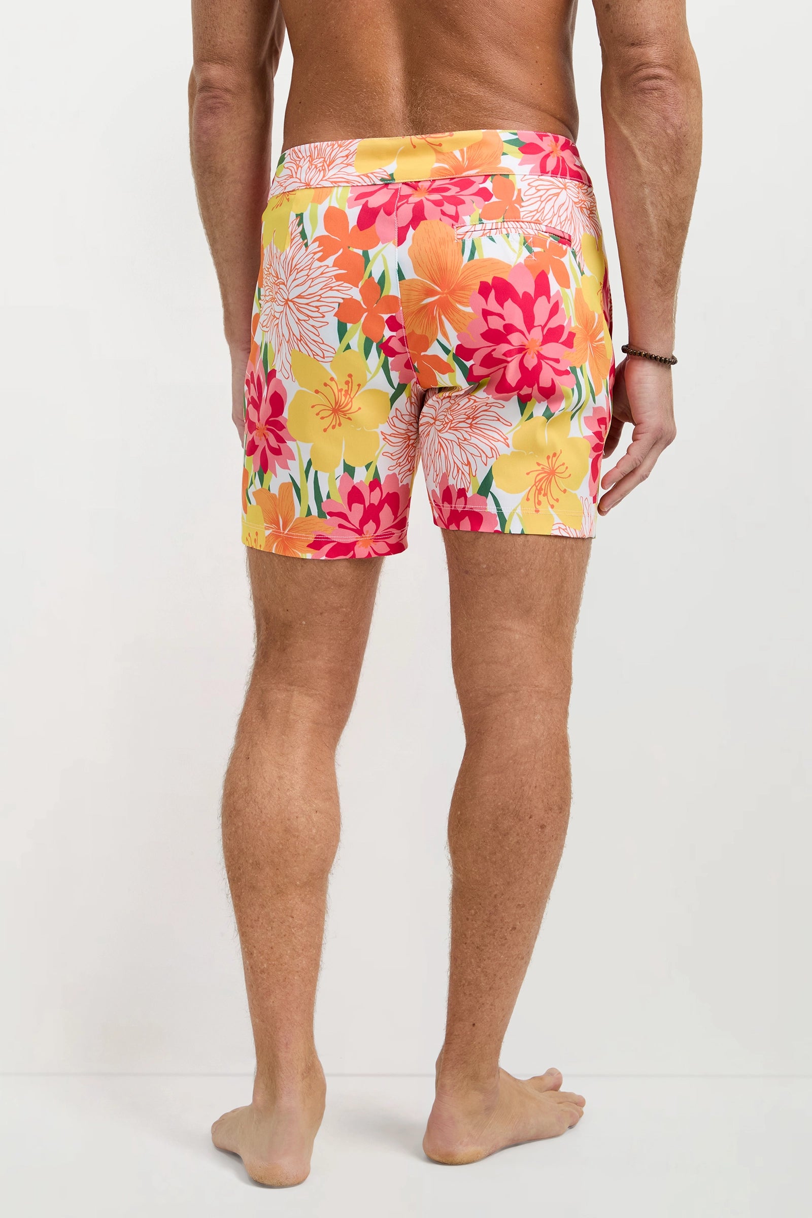 A man stands barefoot, facing away, wearing the Honolulu Board Short - Como featuring a vibrant floral pattern in yellow, orange, pink, and red on a light background.