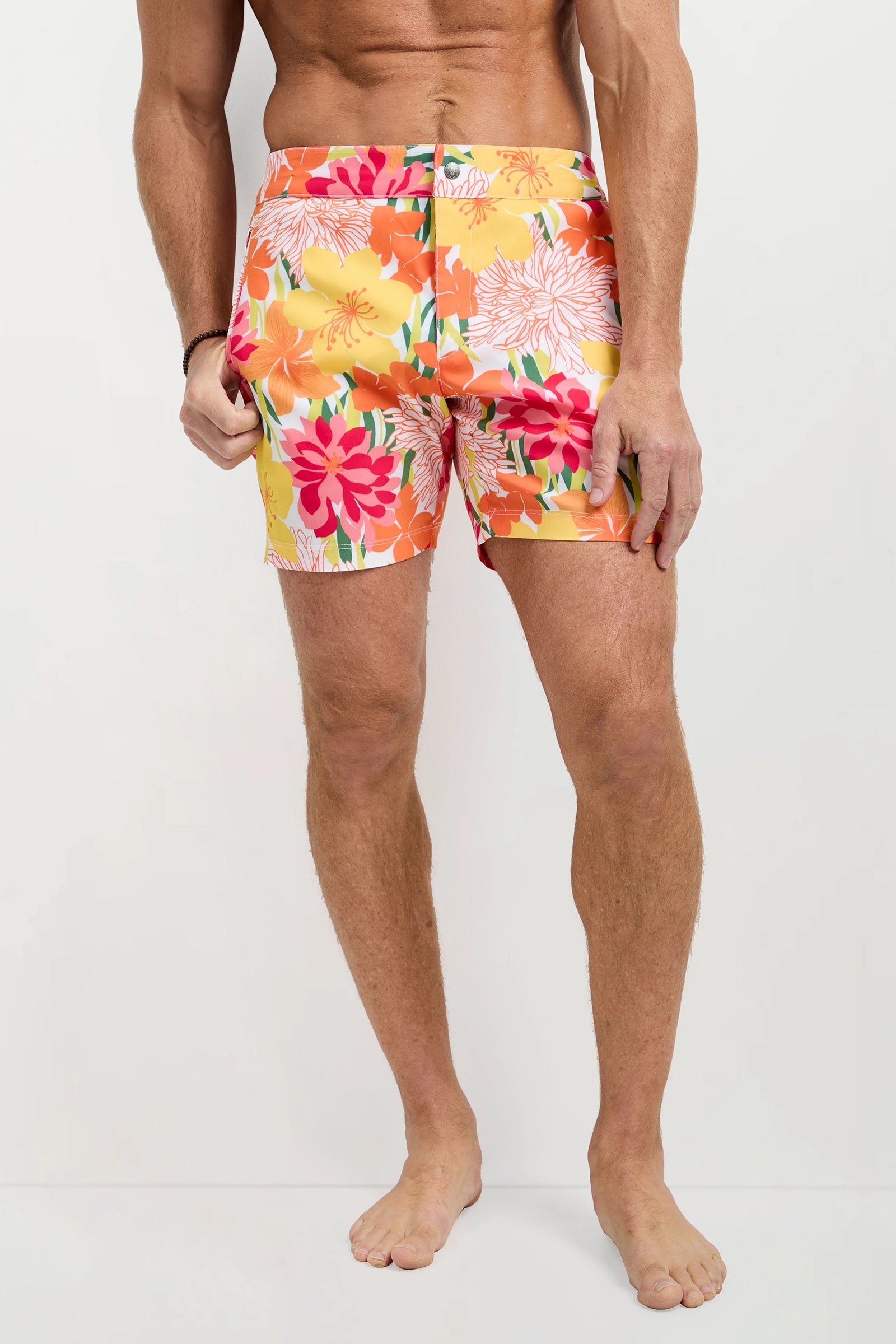 A man stands barefoot against a plain background wearing the Honolulu Board Short - Como, featuring bright floral prints with red, orange, pink, yellow flowers and green leaves, his upper body bare as he holds the waistband with one hand.