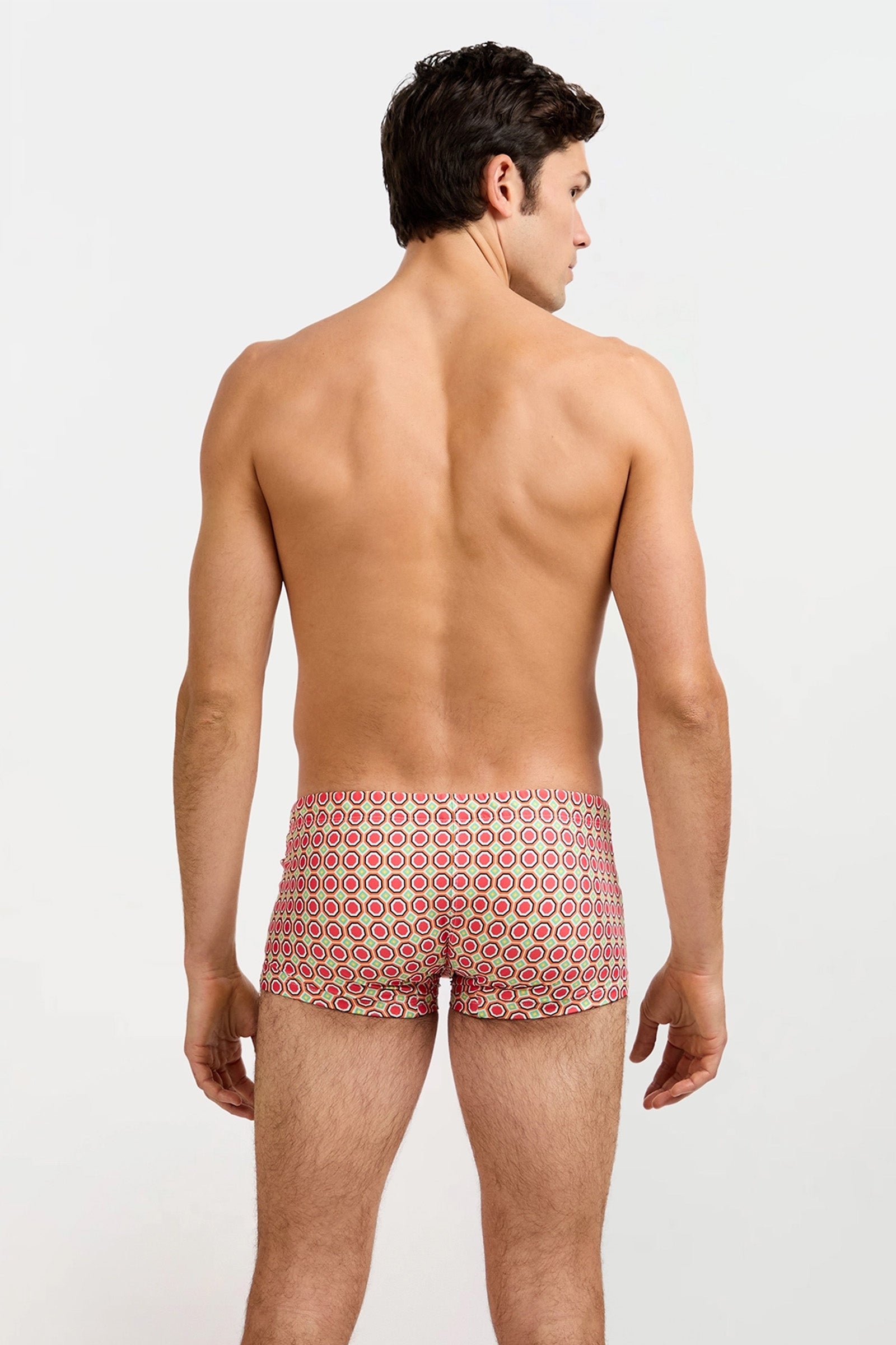 A man, shirtless and facing away from the camera, models the Montego Bay Swim Trunk - Salerno with a red and white circular pattern against a plain light backdrop.