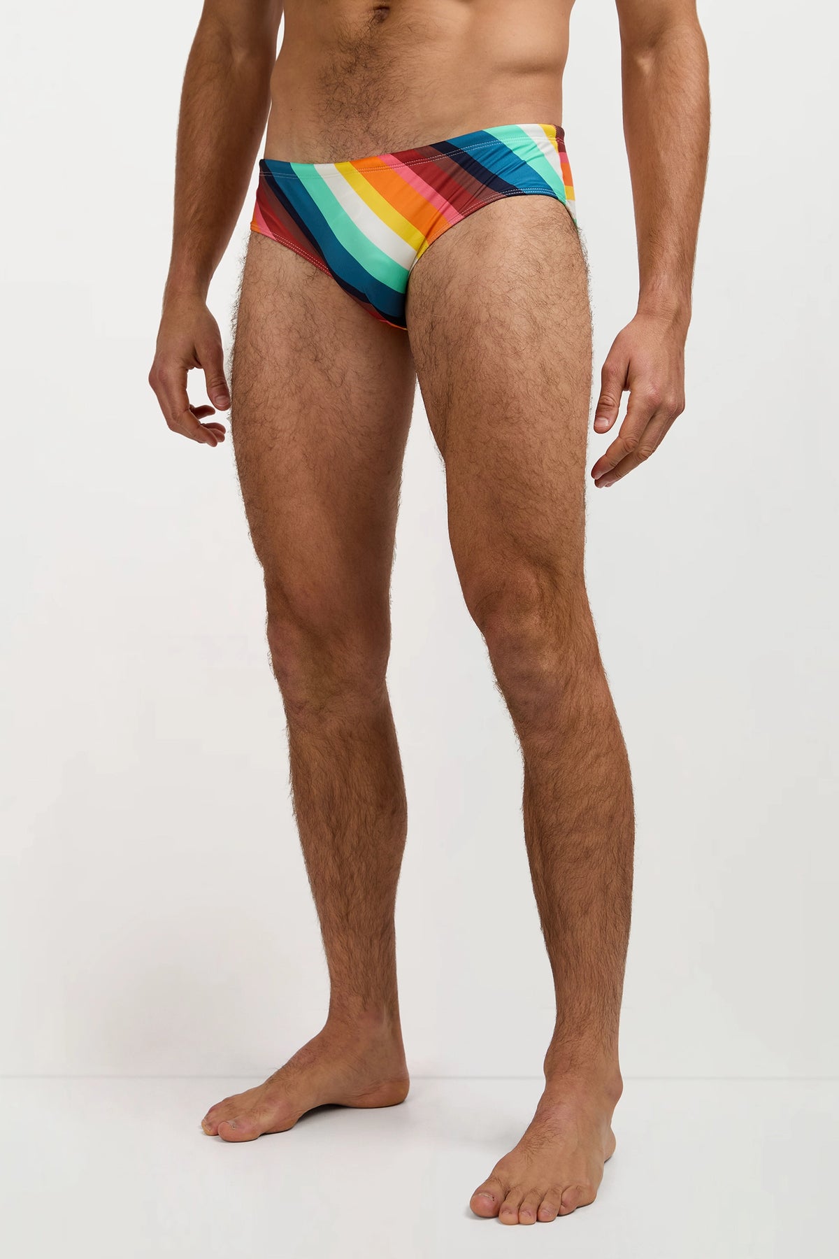 A man stands barefoot, wearing the Bondi Swim Brief - Siena Stripe, which features colorful diagonal stripes. The photo captures his body from shoulders to feet against a plain background.