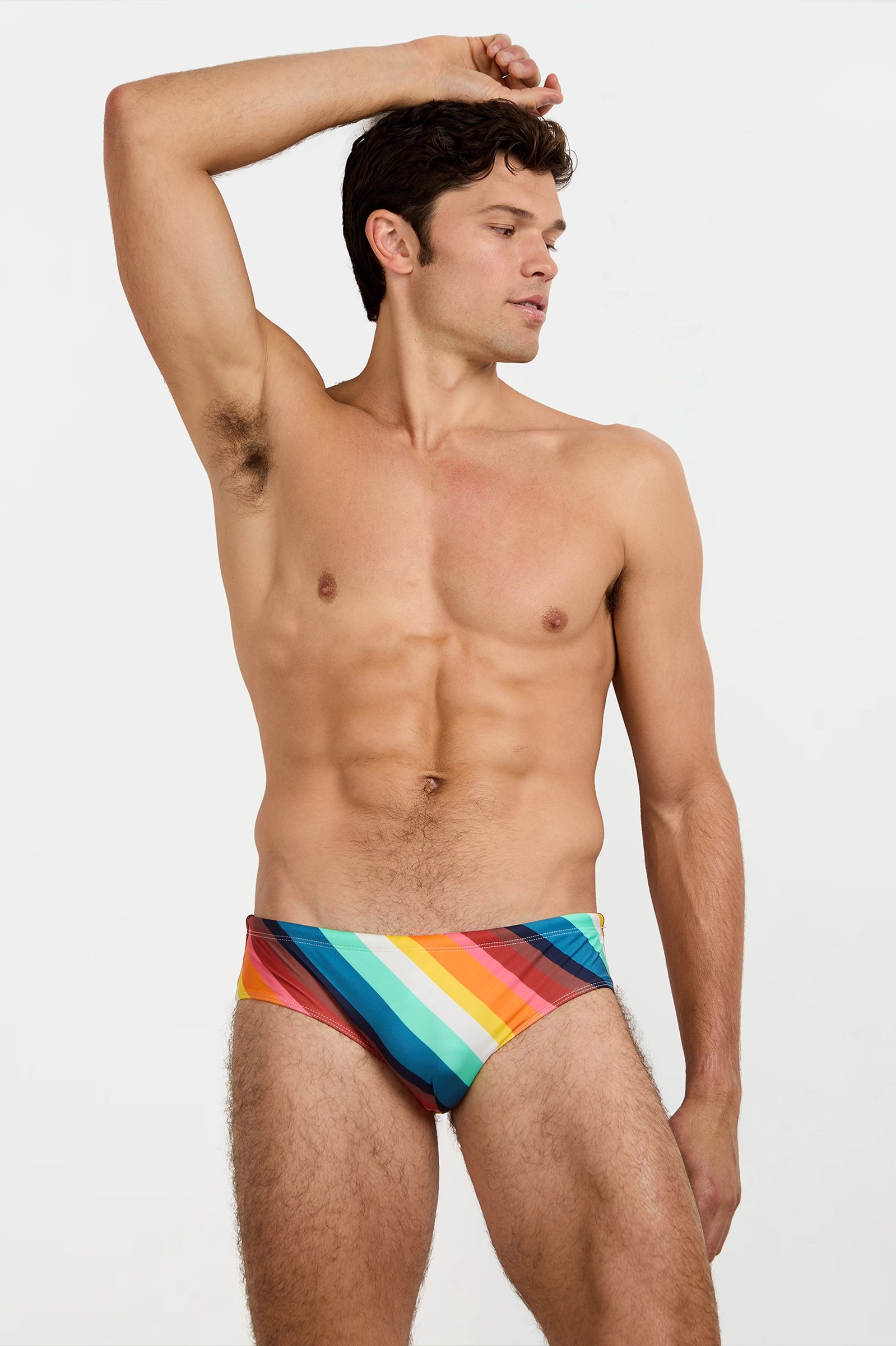 A shirtless man stands with one arm raised, wearing the Bondi Swim Brief - Siena Stripe, featuring colorful rainbow stripes, against a plain white background.