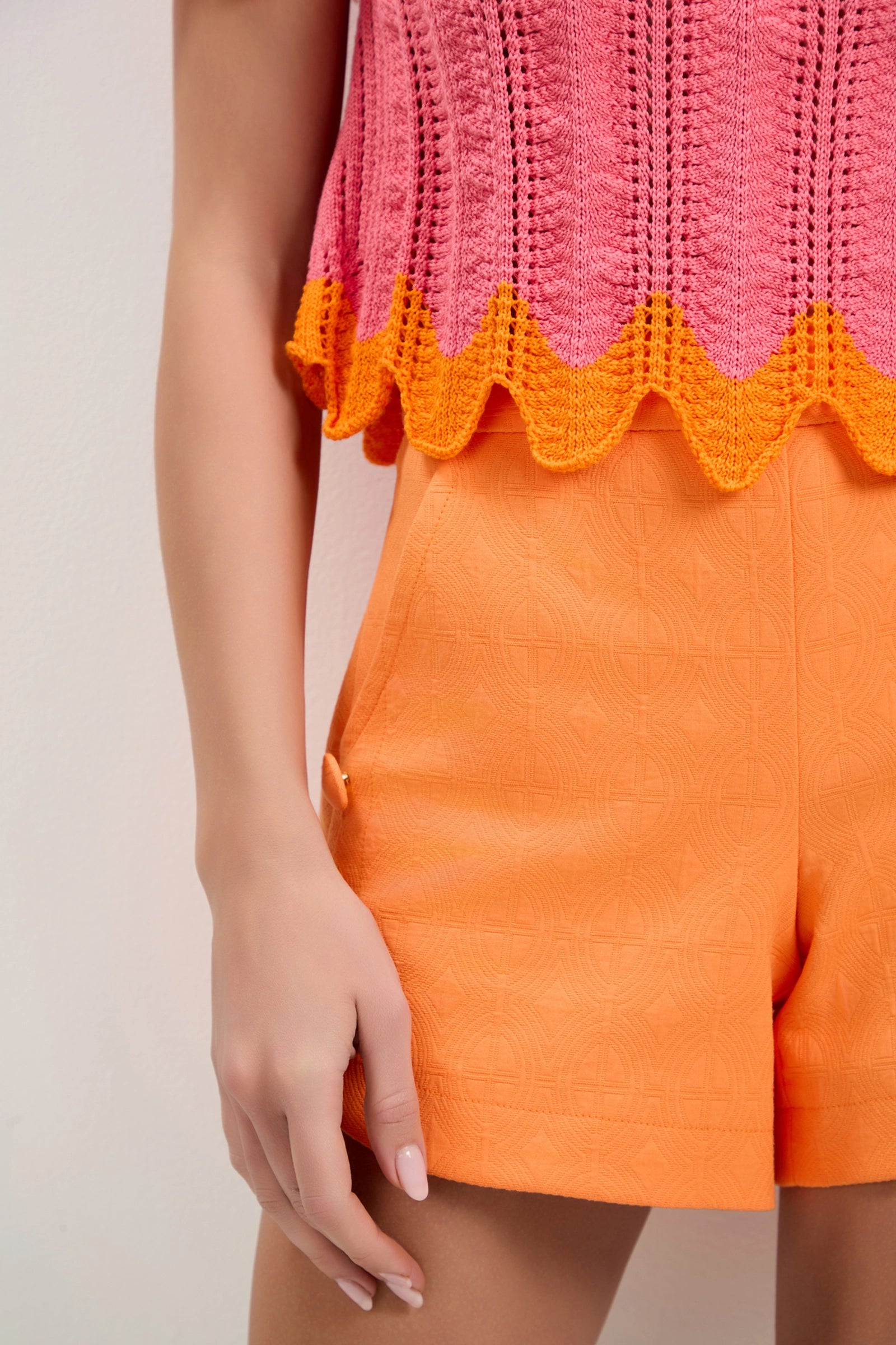A person wearing the Playa Largo Wavy Knit Top in pink with an orange scalloped hem and textured orange shorts, standing against a plain light background. Only their torso and upper legs are visible.