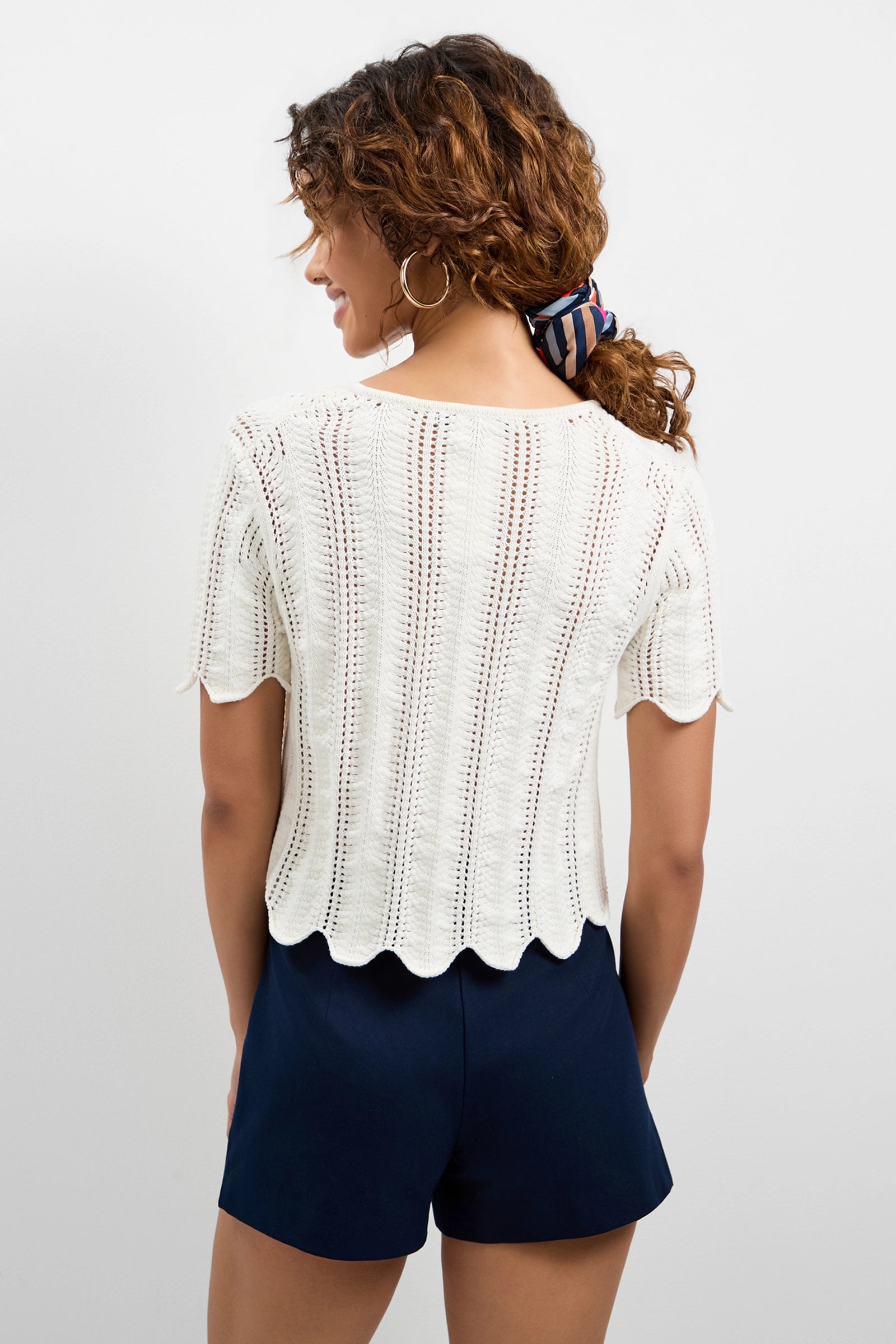 A woman with curly hair in a striped scrunchie is seen from the back wearing the Playa Largo Wavy Knit Top, a white short-sleeve knit with a scalloped hem, paired with navy blue shorts.