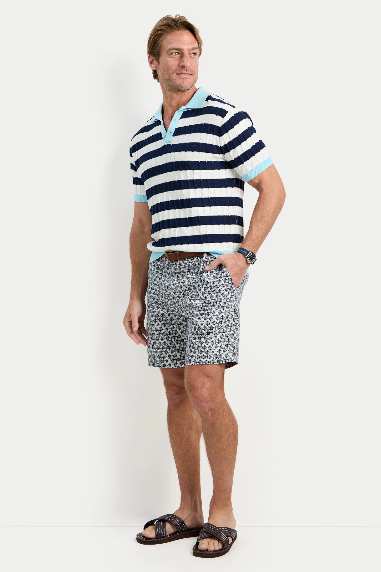 A man wearing the Enzo Short-Sleeve Polo in navy and white stripes, gray patterned shorts, a brown belt, black sandals, and a watch stands against a plain white background.