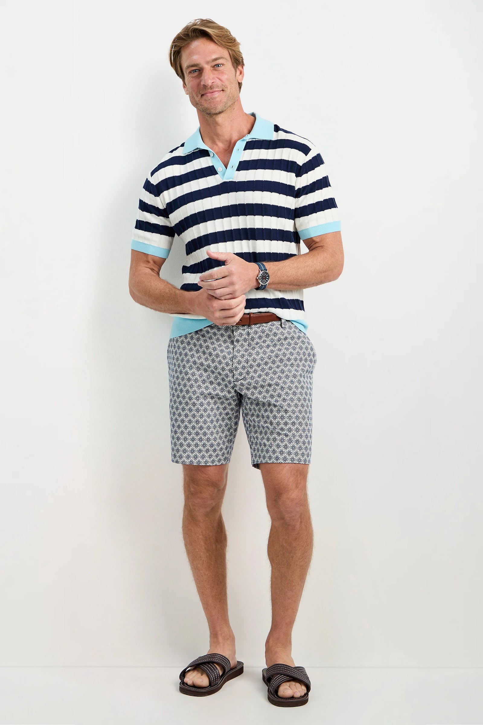 A man smiles against a plain background, wearing the Enzo Short-Sleeve Polo in navy and white stripes, patterned gray shorts, a brown belt, dark sandals, and a watch.
