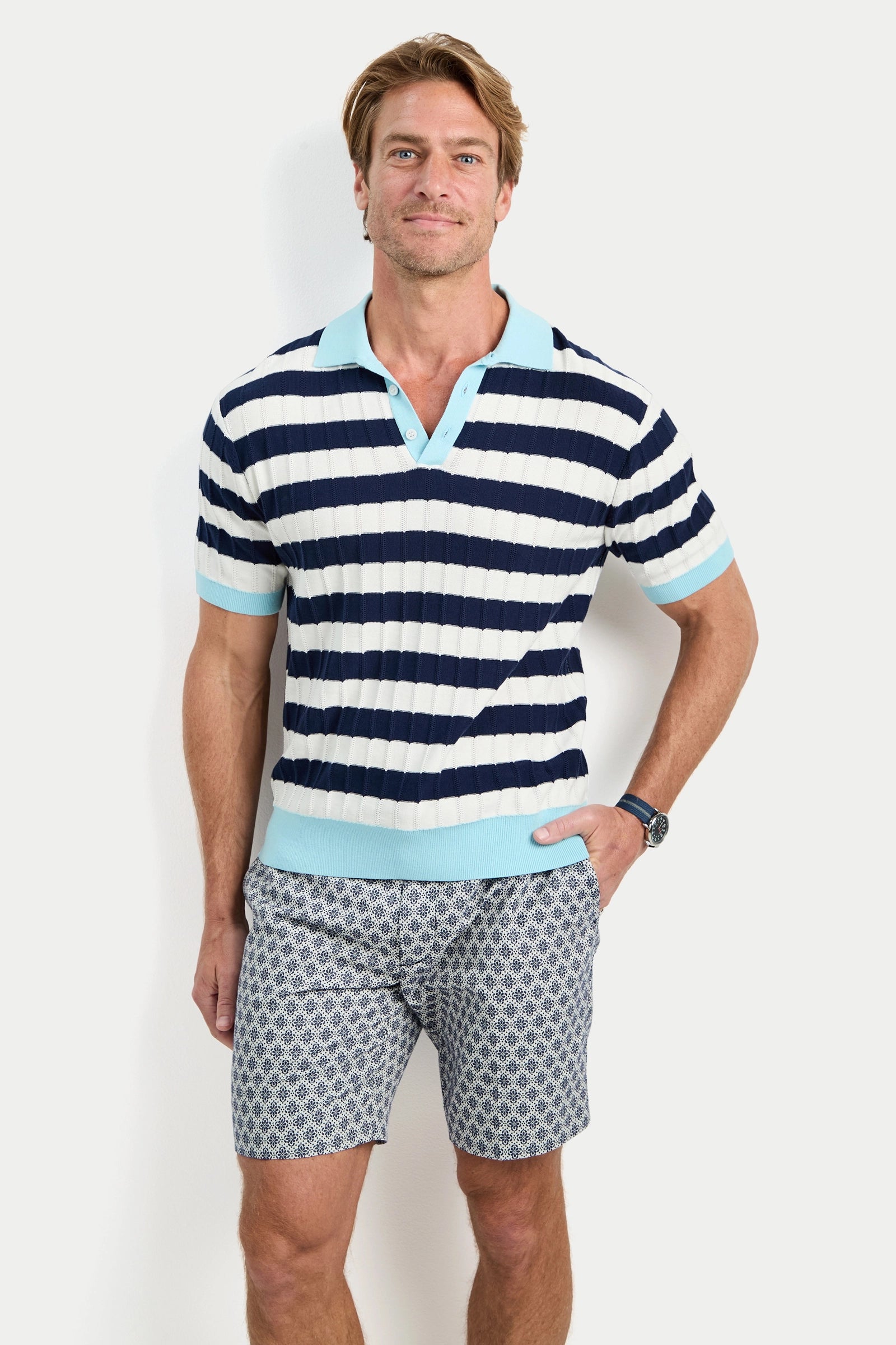 A man stands against a white background, wearing the Enzo Short-Sleeve Polo in blue and white stripes with patterned shorts. He smiles slightly with one hand in his shorts pocket.
