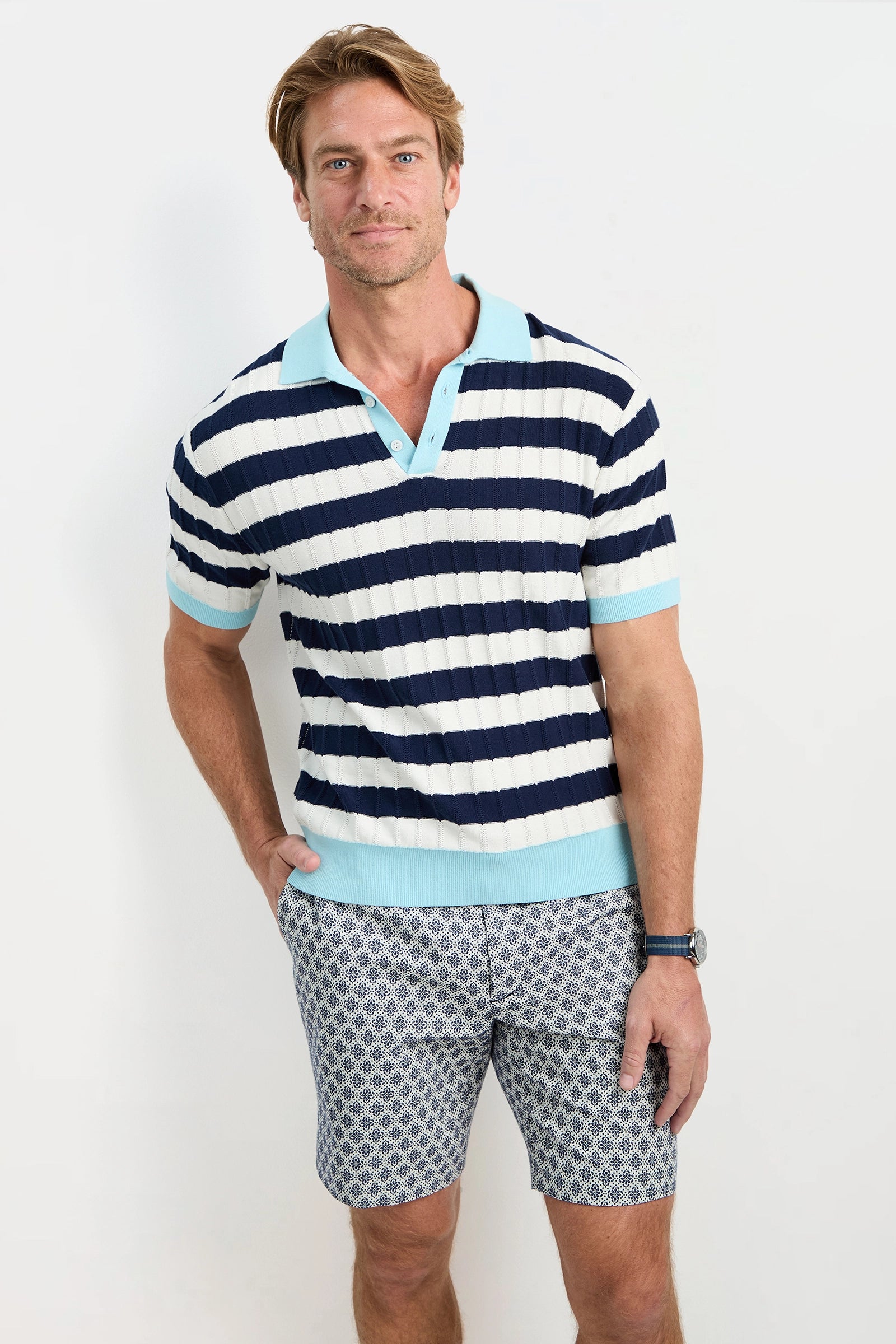 A man with light brown hair and a neutral expression stands against a white background wearing the Enzo Short-Sleeve Polo, featuring navy blue and white stripes, a light blue collar and sleeve hems, paired with patterned blue shorts.