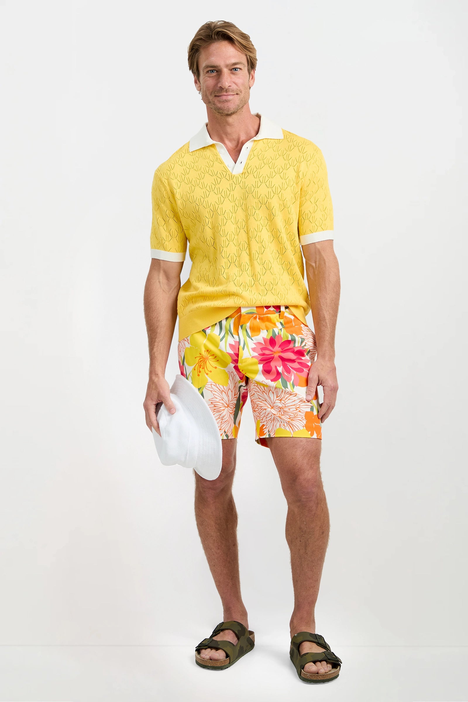 A man smiles at the camera against a white background, wearing the yellow Luca Short-Sleeve Polo, colorful floral shorts, green sandals, and holding a white hat in his right hand.