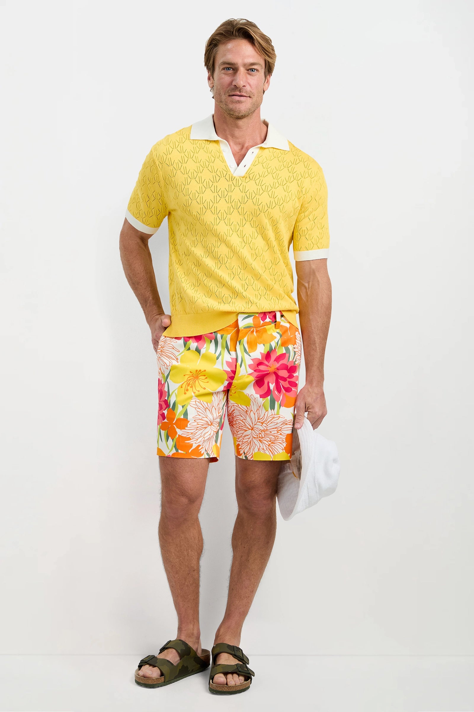 A man stands against a white background wearing Lawrence Short - Como Herringbone, a yellow patterned polo shirt, dark sandals, and holding a white cap by his side.