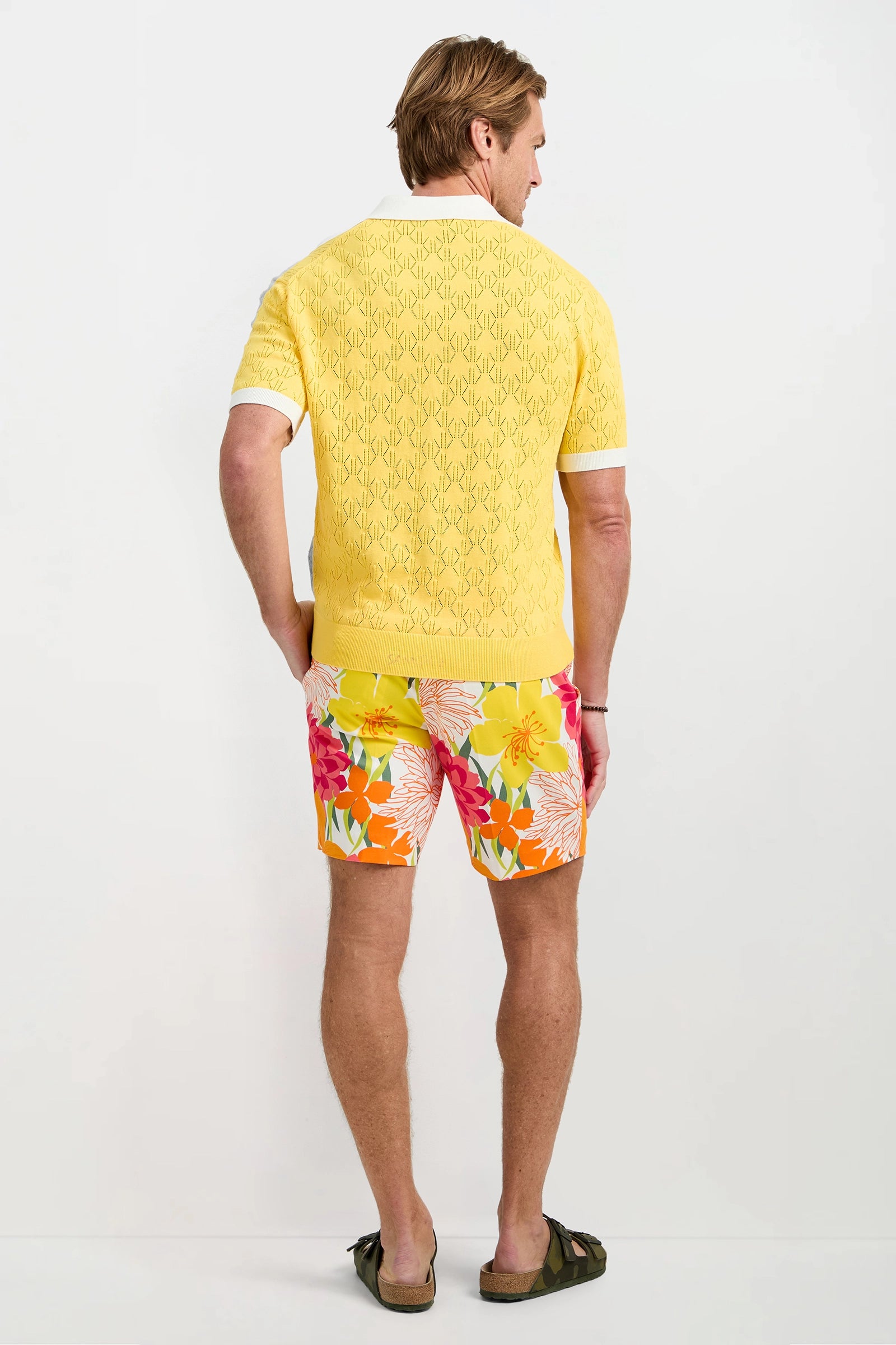 A man faces away, wearing the Luca Short-Sleeve Polo in yellow with a repeating pattern, paired with colorful floral shorts and green sandals, set against a plain white background.