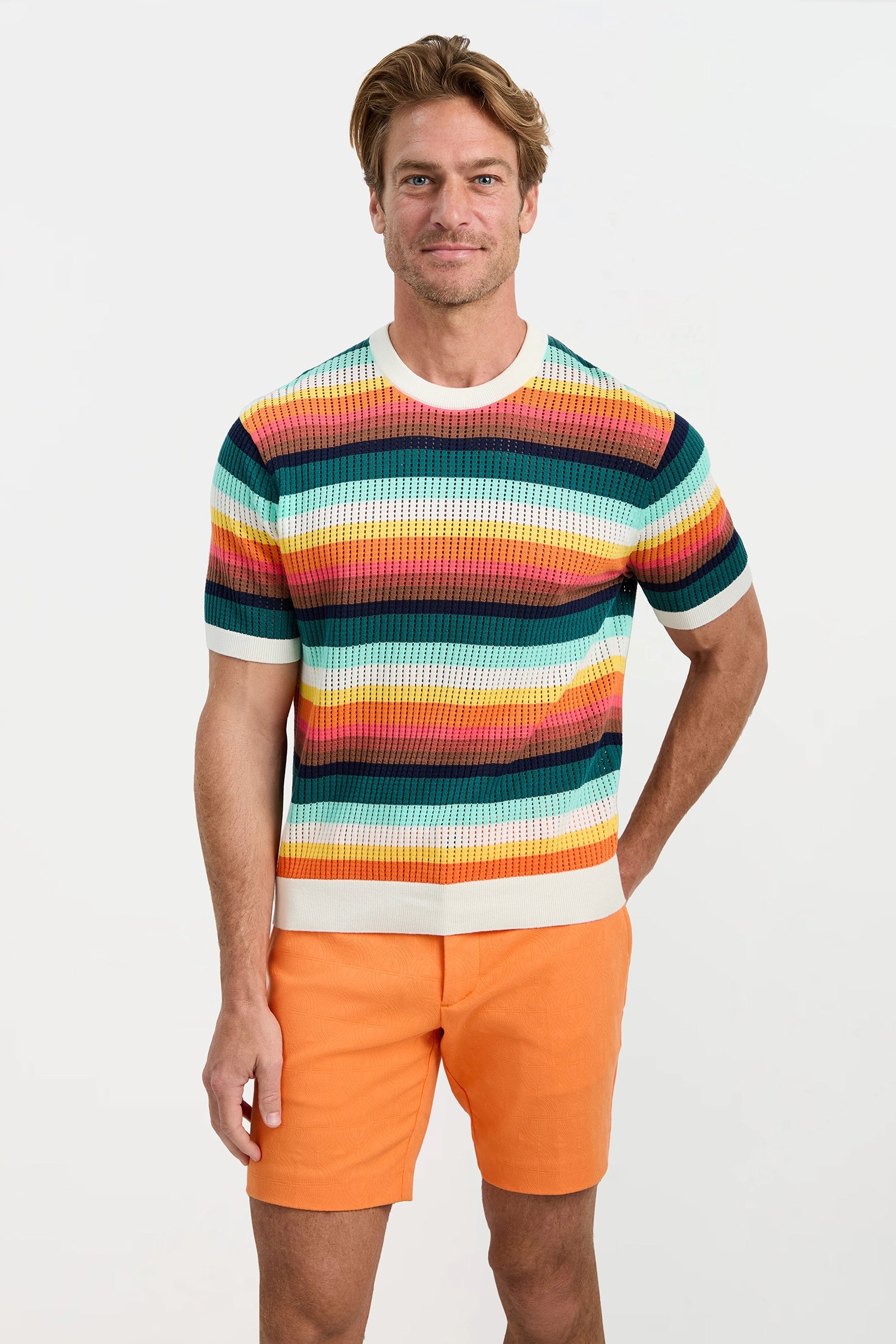 A man stands against a plain white background, smiling as he wears the Carlos Short-Sleeve Mesh Crewneck in colorful stripes with bright orange shorts. One hand rests on his hip while the other is by his side.