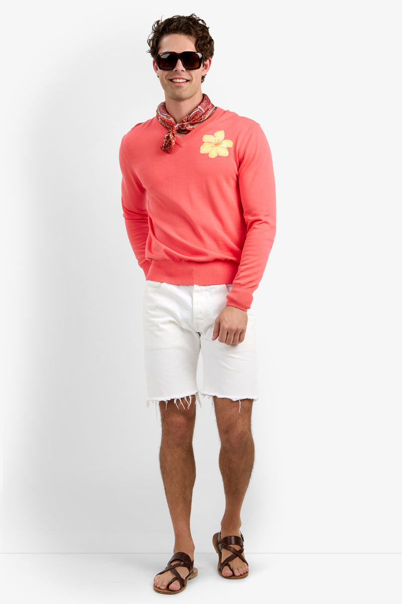 A man in a Divino V-Neck Sweater with a yellow flower patch, white frayed shorts, brown sandals, sunglasses, and a red patterned scarf stands smiling against a white background.
