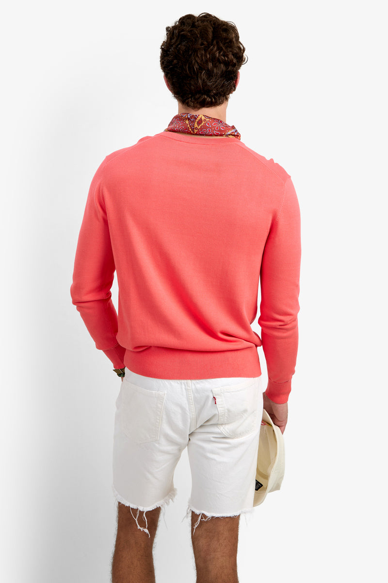 A man, shown from behind against a white background, wears the Divino V-Neck Sweater in coral, paired with white frayed shorts and a patterned neck scarf, holding a light hat in his left hand.